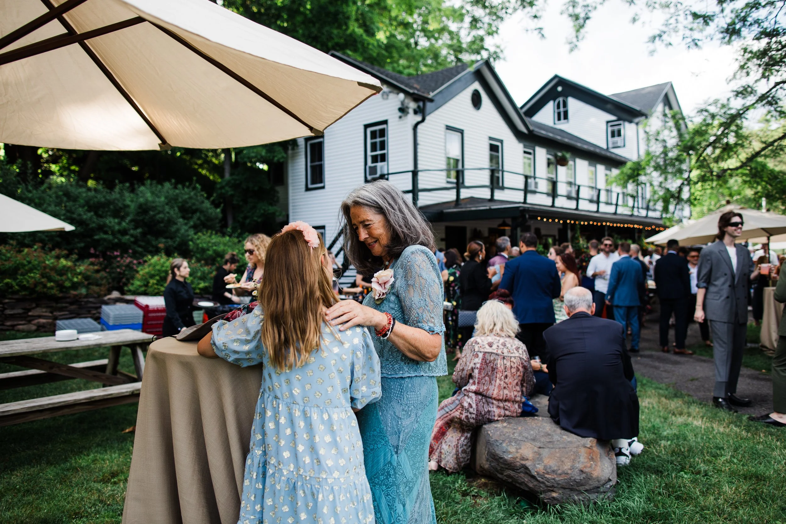 People enjoying outdoor wedding reception at a house with white siding, surrounded by greenery and large umbrellas for shade.