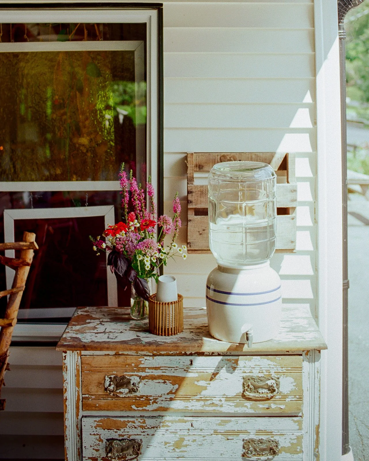 A vintage wooden dresser with peeling white paint, on top of which sits a glass water dispenser, a small vase of colorful wildflowers, and a wicker cup holder with white cups. Behind, part of a house with white siding and a sliding glass door is visi
