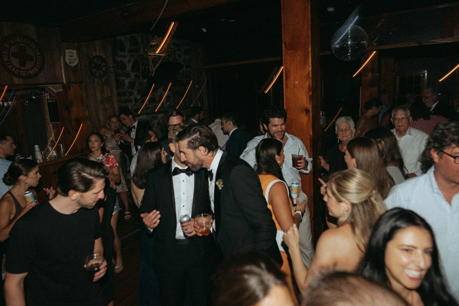 People dancing and socializing at a party or wedding reception in a dimly lit rustic venue with wooden walls and a disco ball.