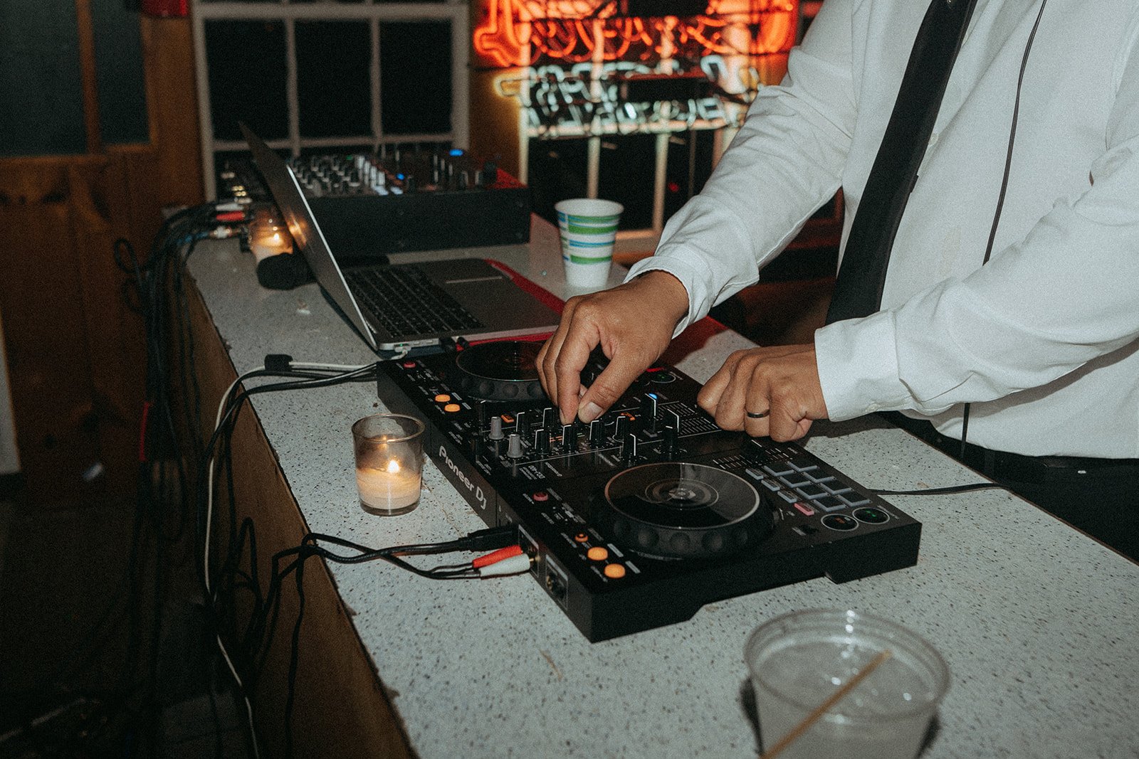 DJ performing with equipment on a table, including a laptop and candles, at a dimly lit venue with neon signs in the background.