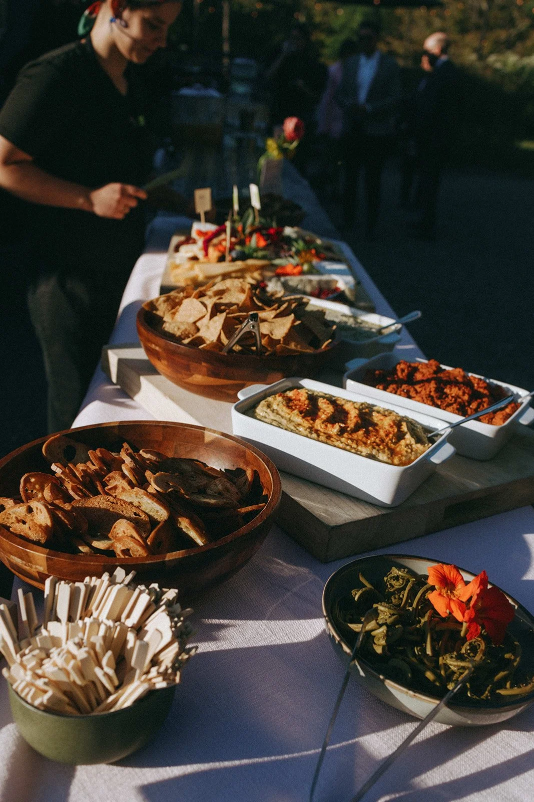 A buffet-style outdoor table with various dishes including chips, dips, vegetables, and bread. A person is serving themselves, and there are people in the background, some wearing masks.