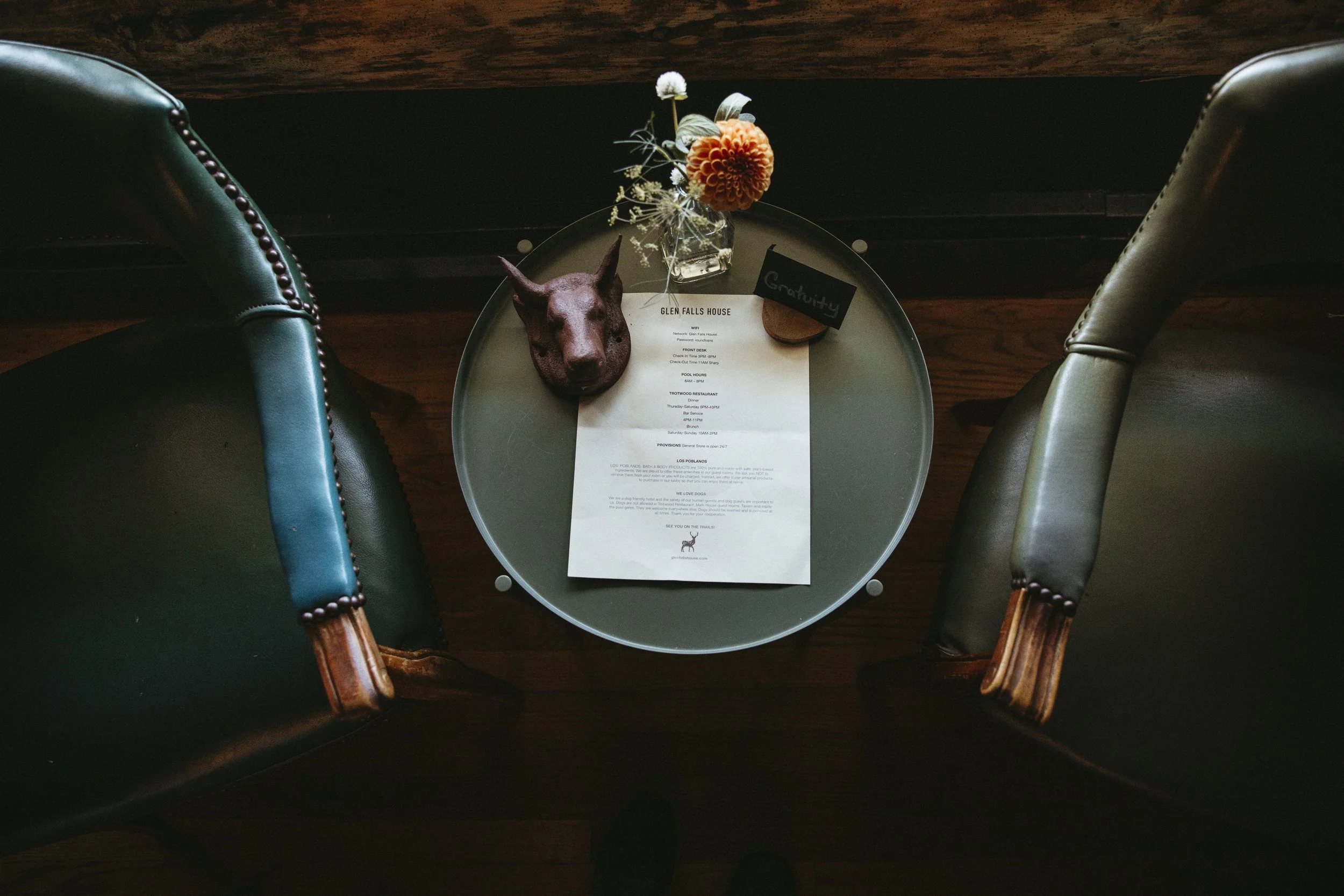A round glass table with a menu, a dog-shaped object, a small sign that says "Gravity," a flower arrangement, and a martini glass, surrounded by two dark green leather chairs on a wooden floor.