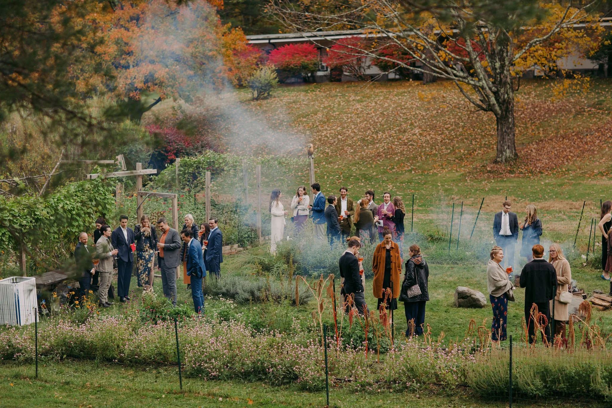 People gathered outdoors in a garden during autumn, with colorful fall foliage, some smoking a barbecue or fire, and dressed in semi-formal attire, engaging in conversations.