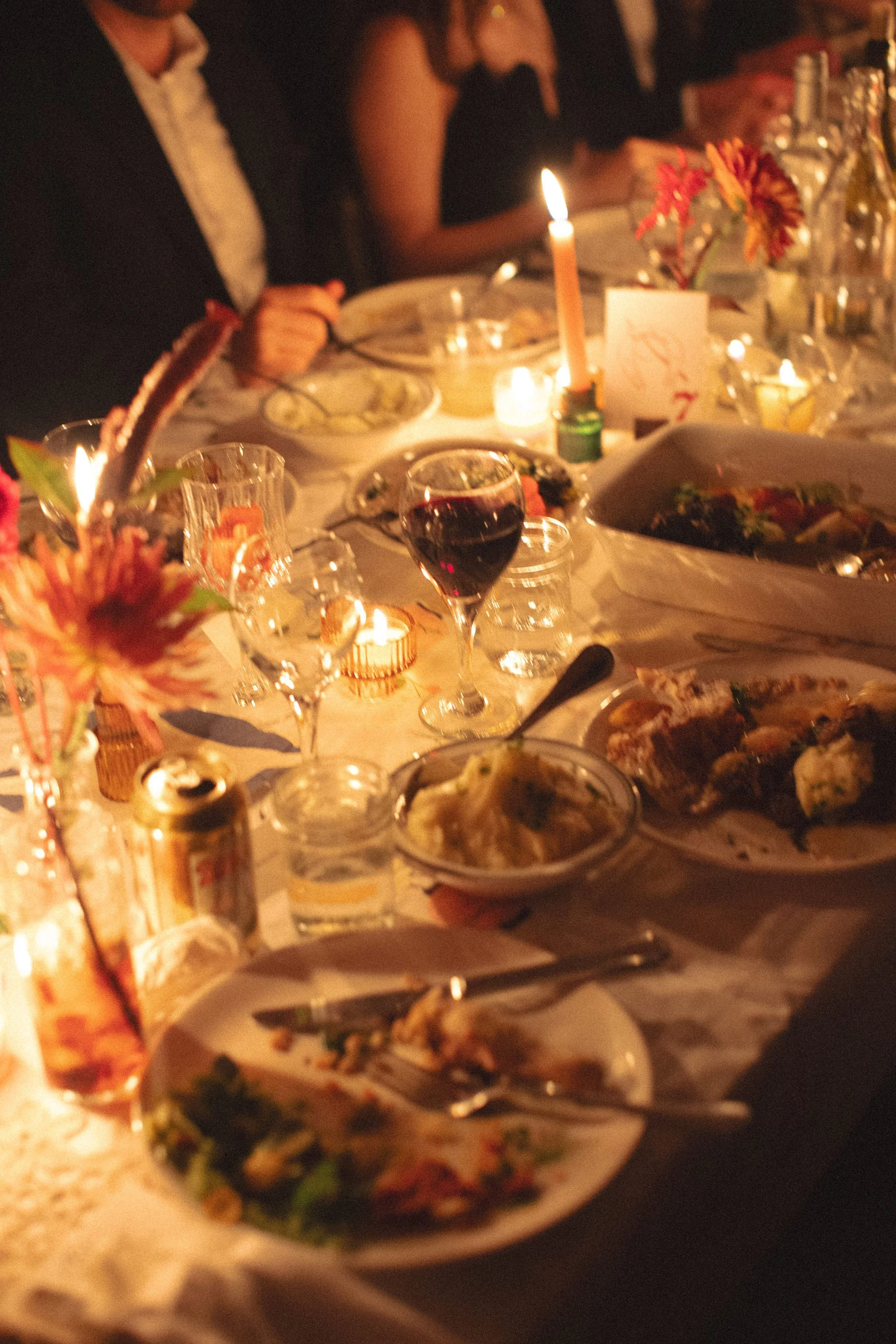 A dinner table set for a celebration, illuminated by candles, with various plates of food, a glass of red wine, and floral centerpieces.