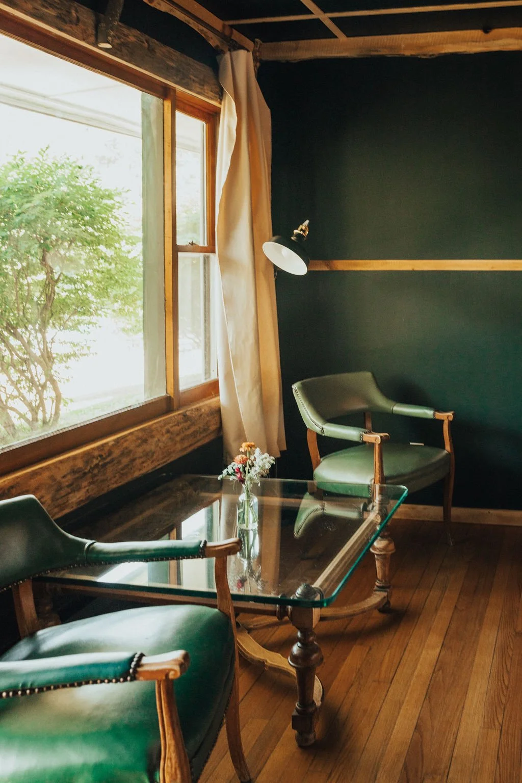 A cozy corner of a room with a glass-top wooden table, two green upholstered chairs with wooden frames, a small vase with flowers, a window with a view of greenery outside, and a wall-mounted lamp.