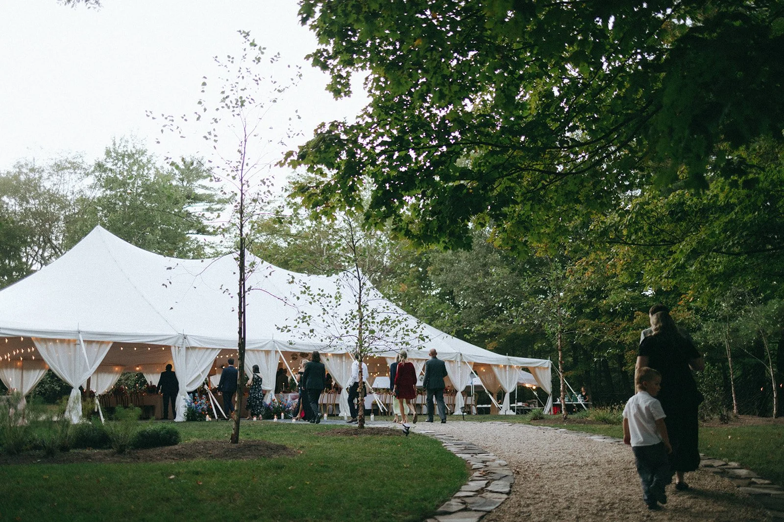 People gather near a large white event tent in an outdoor, wooded area with green trees and a gravel pathway.