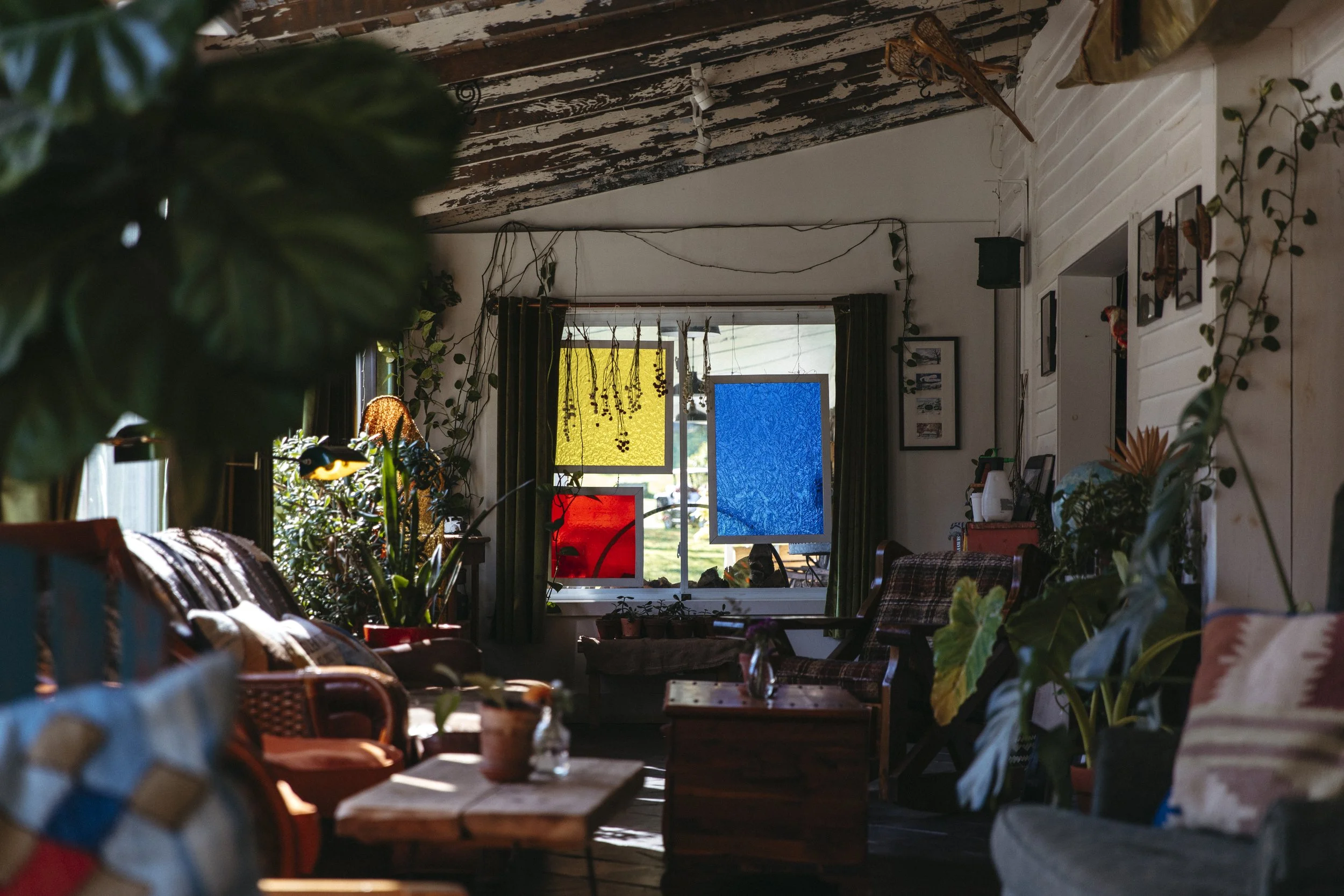 A cozy indoor living room with wooden furniture, plants, and colorful stained glass window panels in blue, red, and yellow.