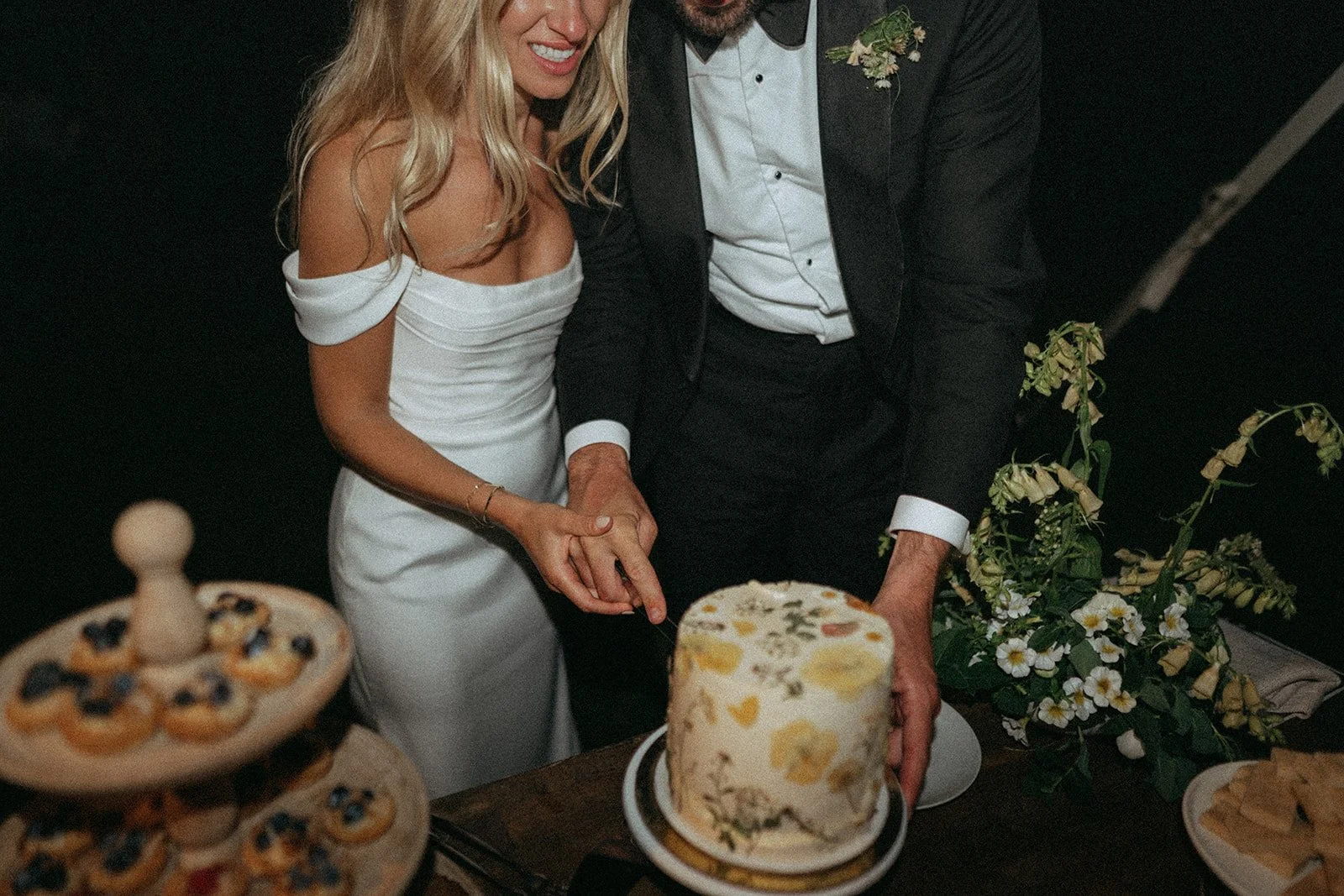 A bride and groom cutting a wedding cake together, with the bride in a white off-the-shoulder dress and the groom in a black tuxedo, surrounded by wedding desserts and decorations.