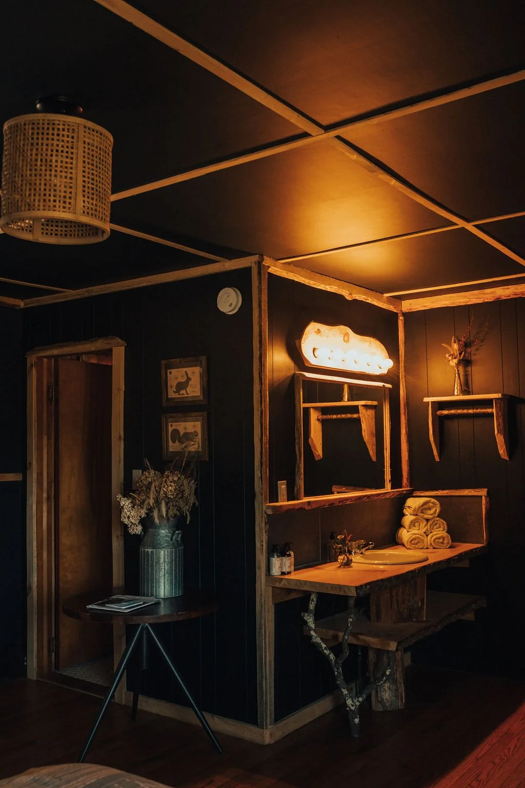 Cozy bathroom with dark wooden walls, warm lighting, rolled towels, a sink, and rustic wooden shelves.