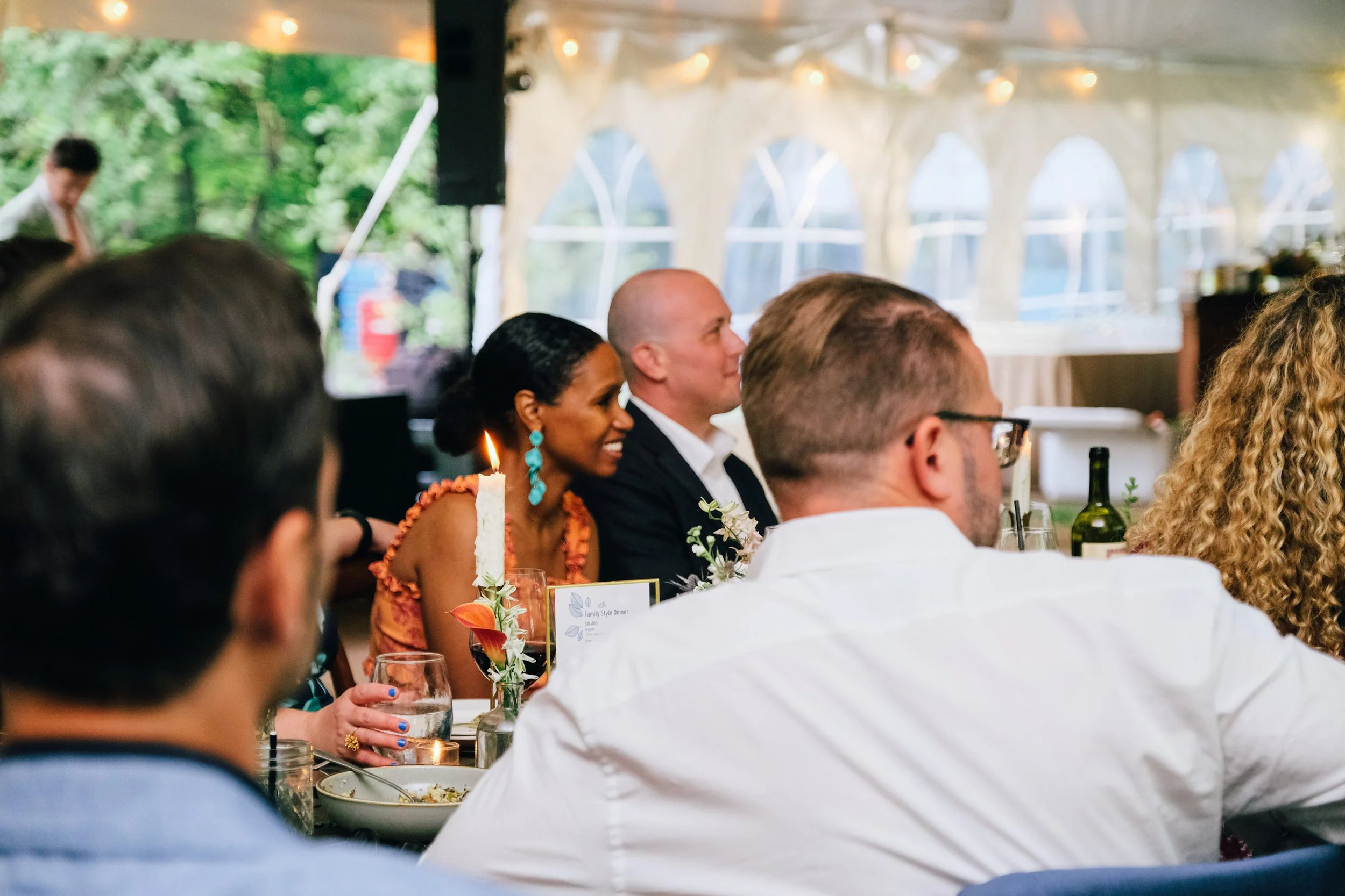 People sitting at a table during an event inside a tent with large windows, enjoying food and drinks, with a woman wearing blue earrings and an orange dress, and a man in a dark suit, in a festive atmosphere.