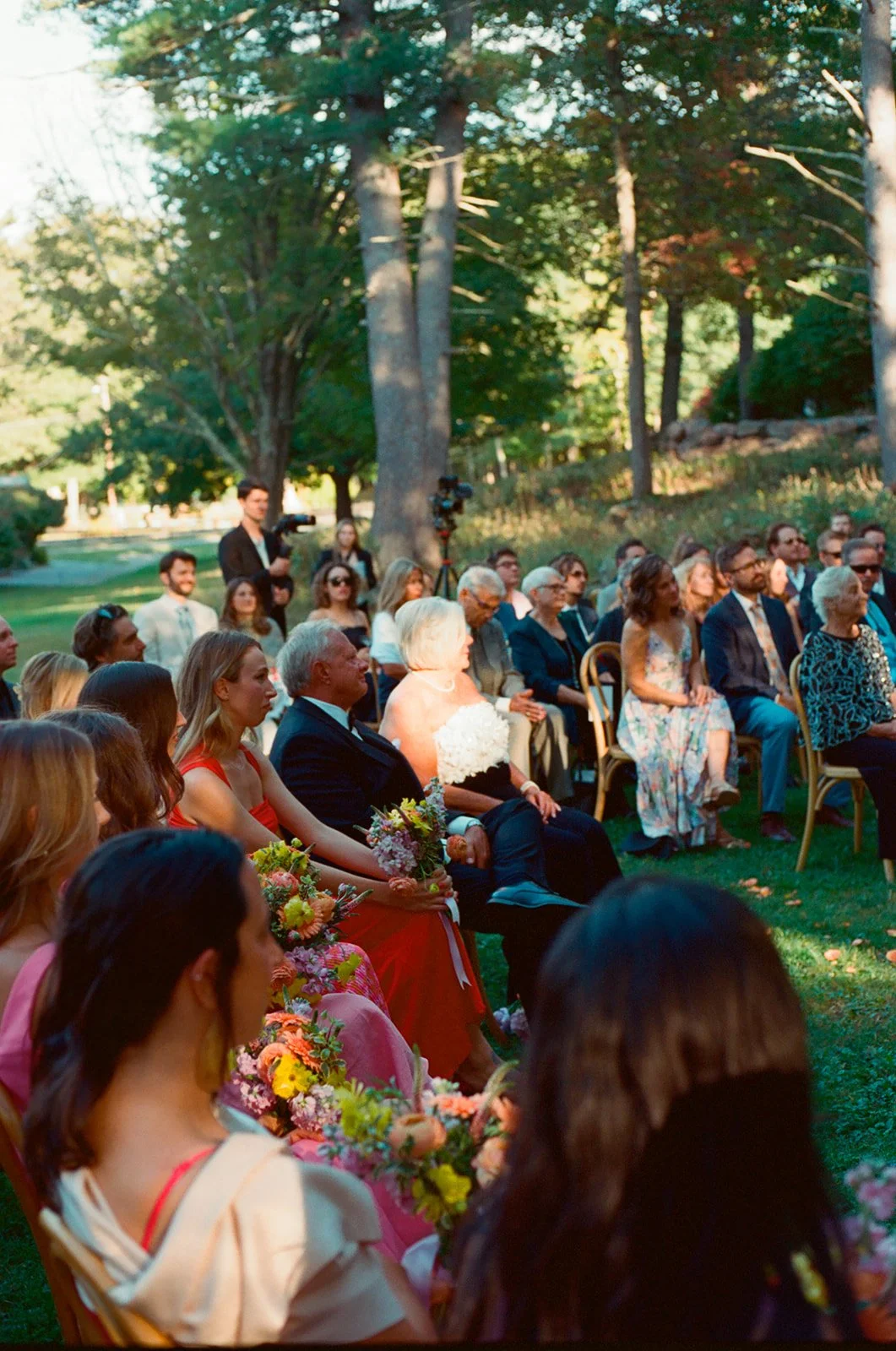 People sitting outdoors at a wedding ceremony, with some holding flower bouquets, surrounded by trees and natural scenery.