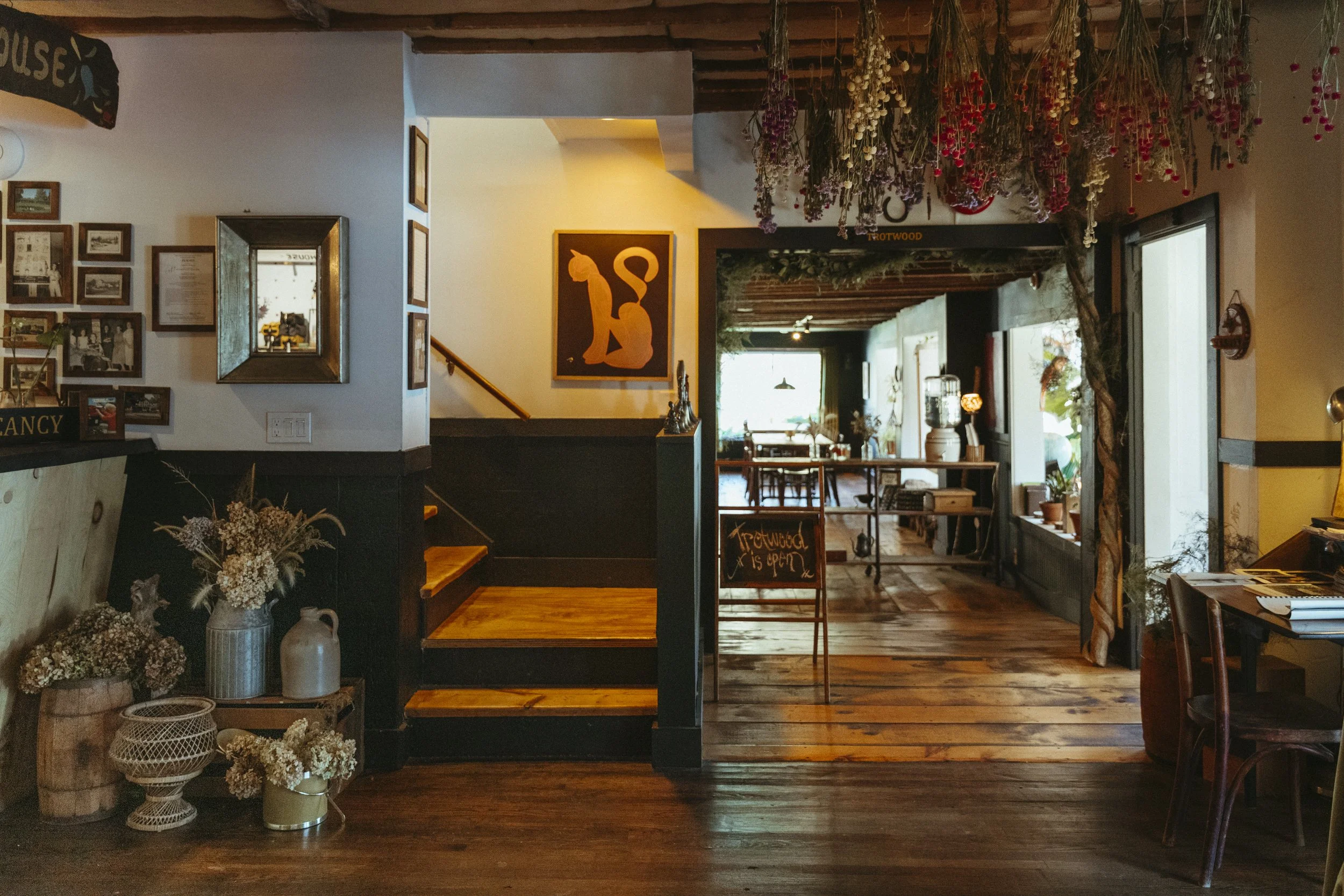 Interior of a cozy cafe or restaurant with wooden floors, decorated with dried flowers hanging from the ceiling, framed photographs and artwork on the walls, and a staircase leading to an upper level. There is a sign on an easel indicating the place 