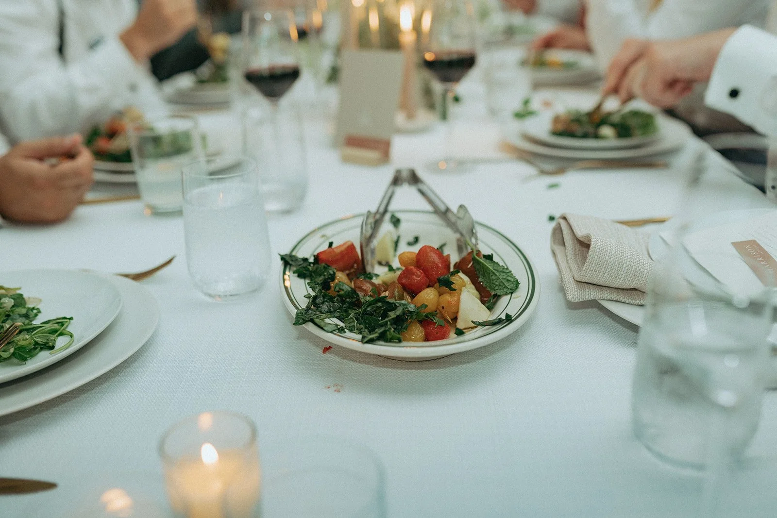 A close-up of a table setting at a formal event with a salad in a white bowl in the center, surrounded by glasses of water and wine, cutlery, and napkins on a white tablecloth.