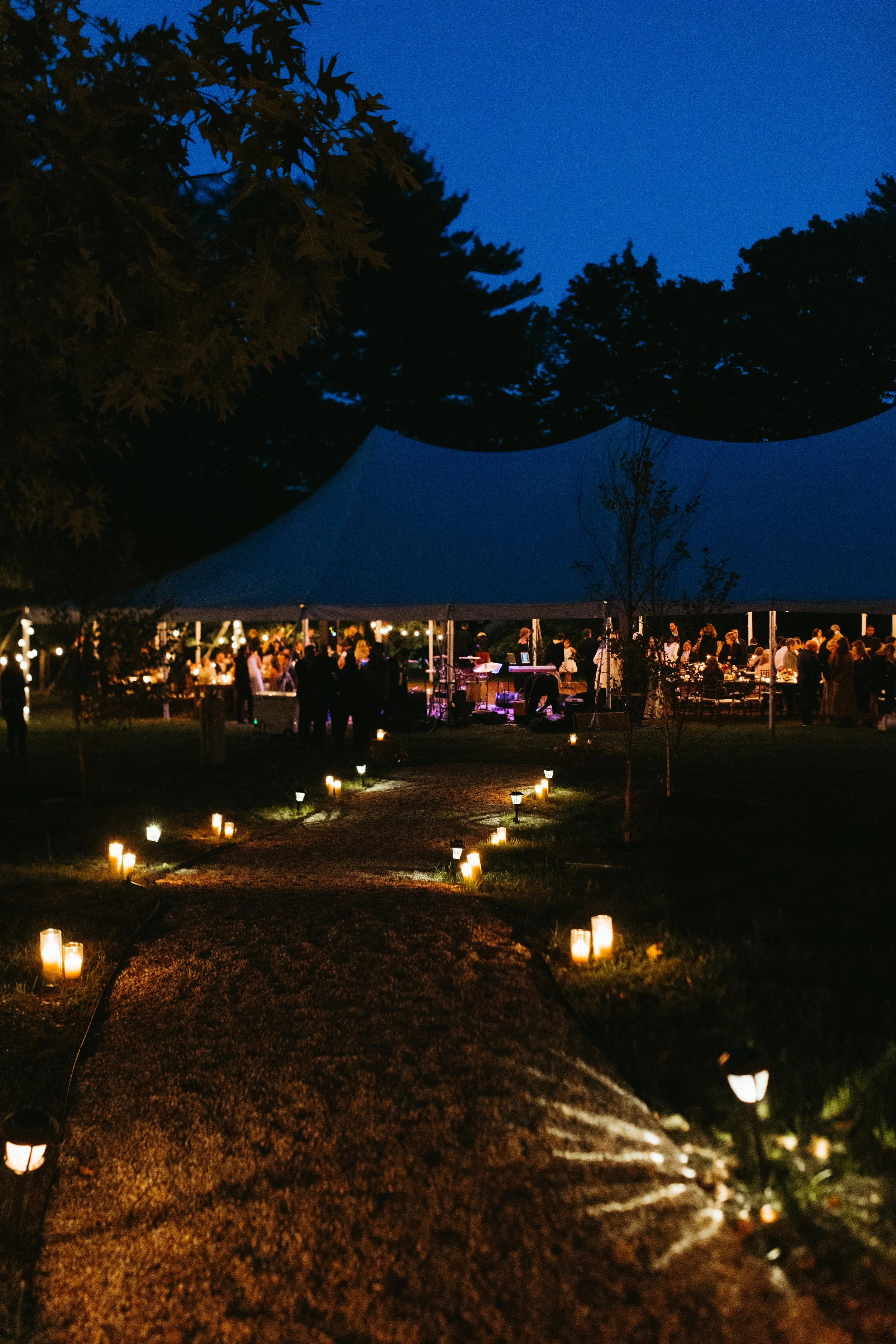 Nighttime outdoor gathering under a large tent with string lights, with guests socializing and a band playing music, illuminated by small pathway lights.