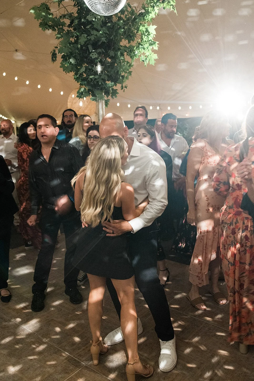 A group of people at a party or dance event, with a couple in the center dancing closely, surrounded by others socializing and dancing, under string lights and a large artificial tree.