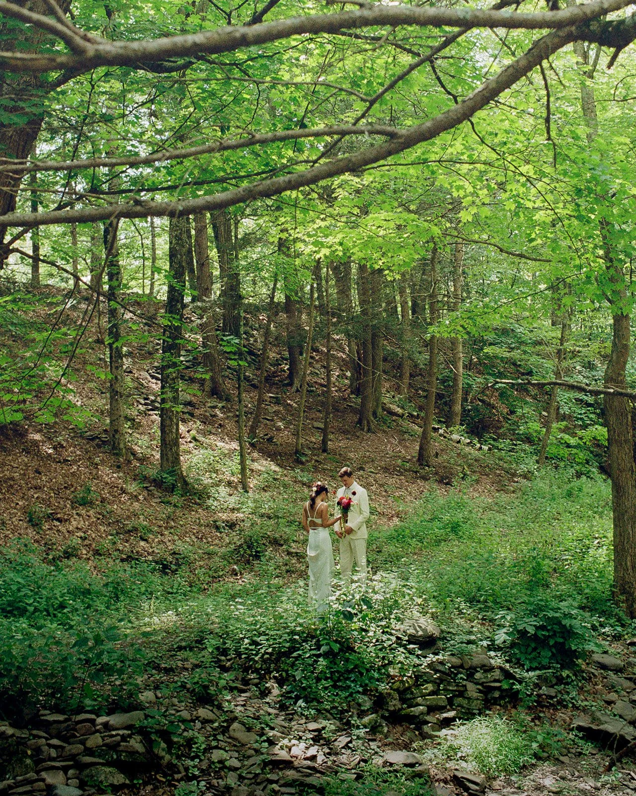 A couple dressed in white, holding flowers, stands on a small stone bridge in a lush green forest, sharing a tender moment.