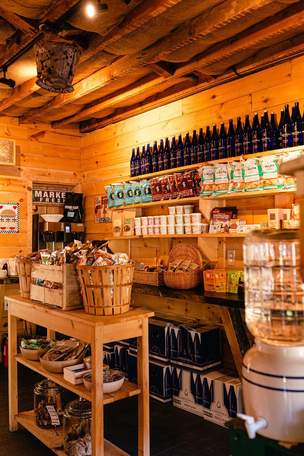 Inside a rustic wooden store with shelves stocked with bottled drinks, snack chips, and other food items, with a wooden table displaying snack baskets and jars of dried goods.