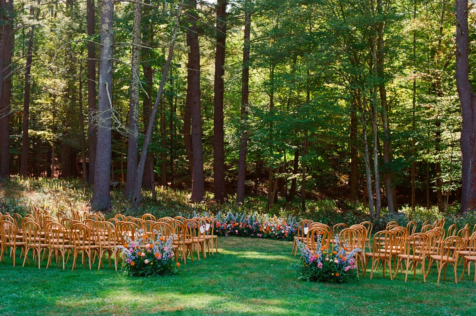 Wedding ceremony setup in a forest with rows of wooden chairs and colorful flower arrangements on a grassy area.