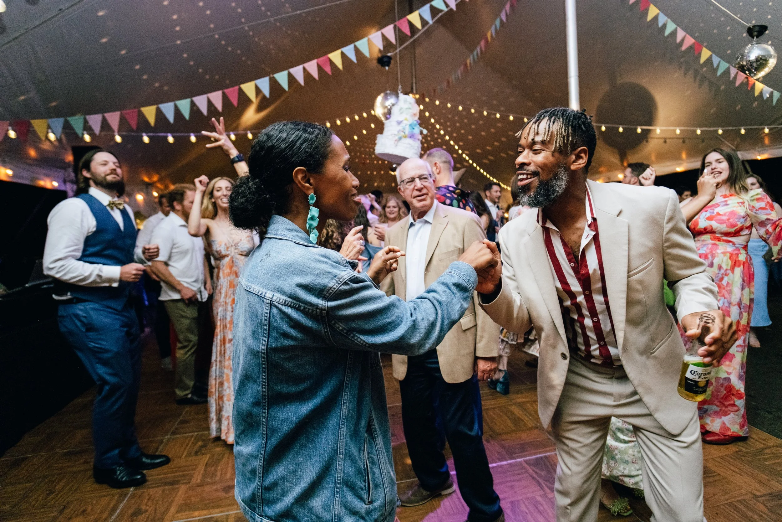 People dancing and socializing at a celebration with colorful banners, string lights, and a cake hanging from the ceiling.