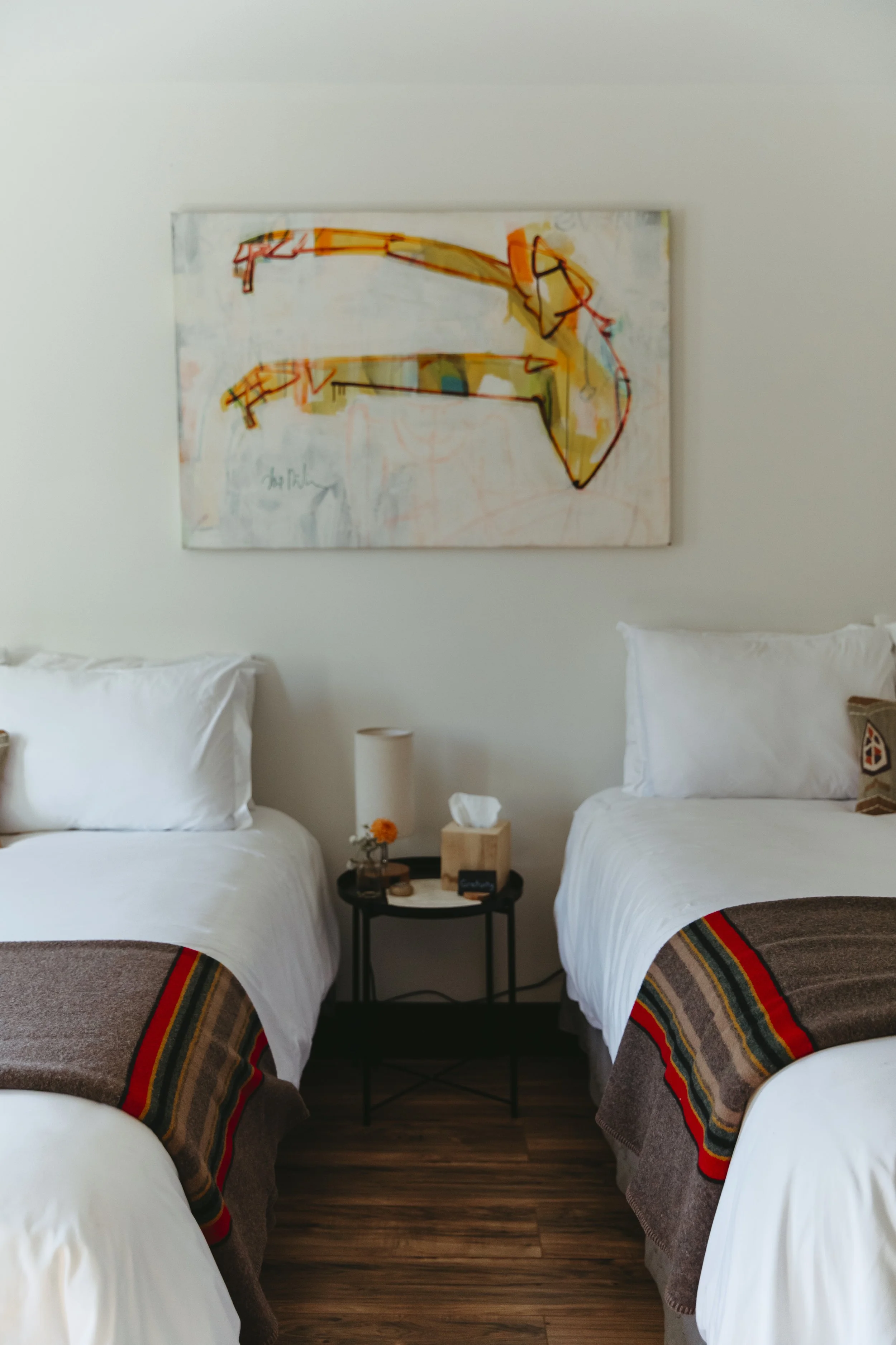 A hotel room with two white beds, a small black side table with a lamp, tissue box, small flower vase, and a hotel sign, with abstract art hanging above the beds.
