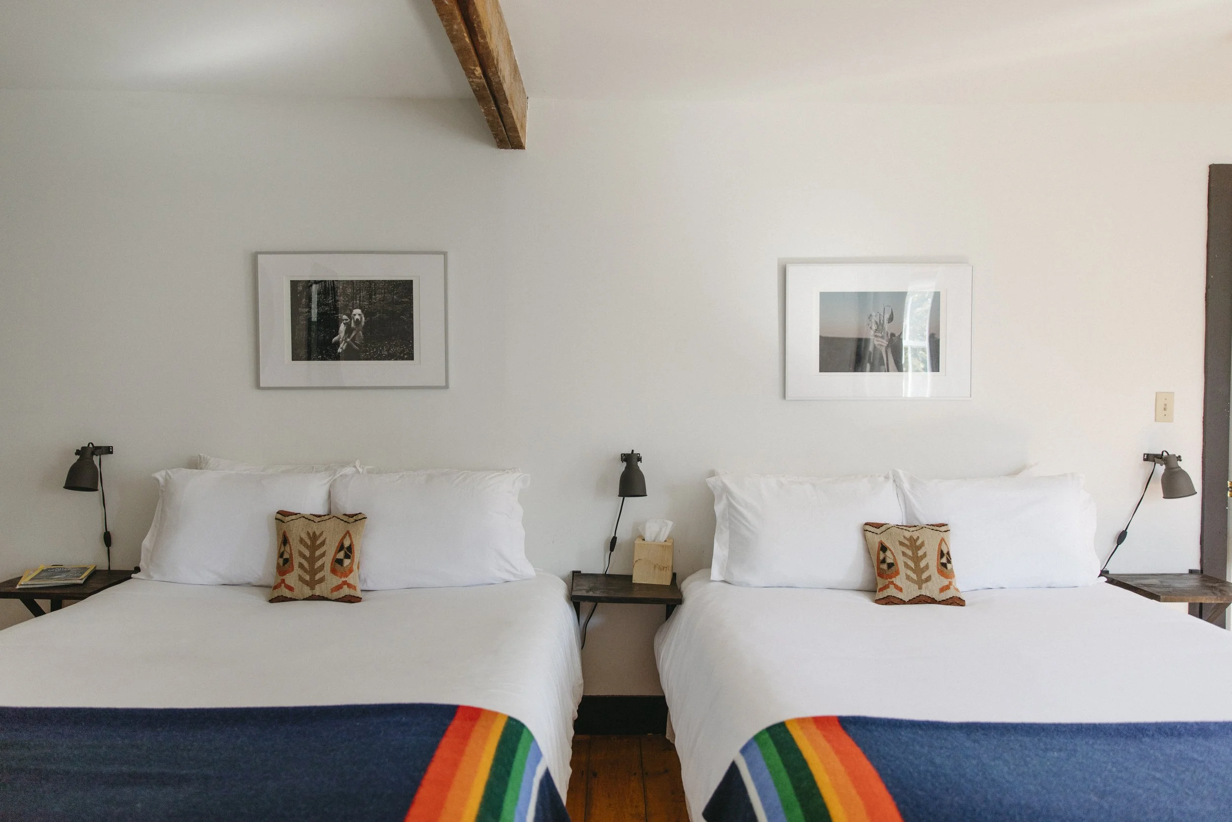 Two beds with white linens and rainbow-striped blankets in a room with white walls, each with small decorative pillows with graphics, black reading lamps, a small wooden nightstand with a tissue box between the beds, and framed black-and-white photog
