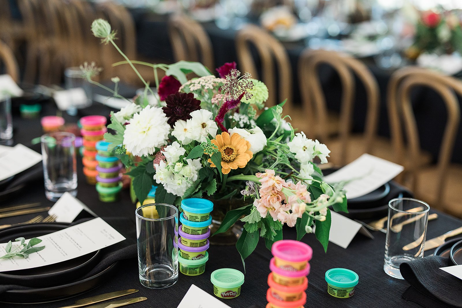 Table decorated with a large floral centerpiece, stacks of Play-Doh containers in various colors, empty glasses, black plates, gold utensils, and printed menus, set for a celebration or event.