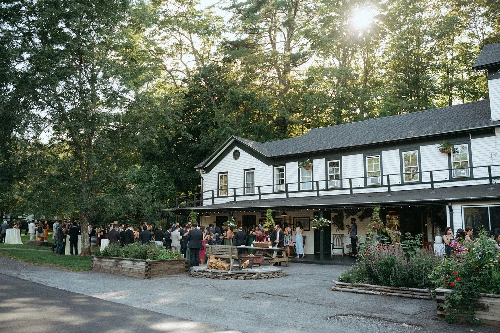 Guests gather outside a two-story white house with black trim for a social event in the evening, with trees in the background and string lights hanging from the porch.