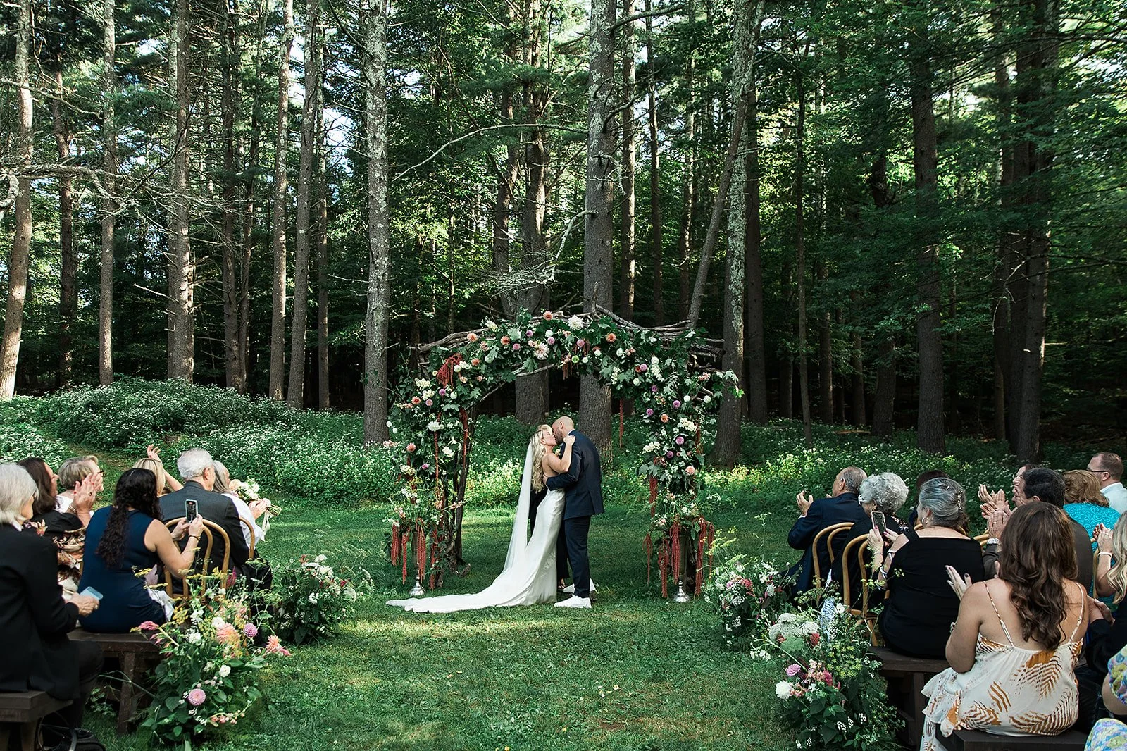 A couple kisses during an outdoor wedding ceremony on a green lawn in a forest, with guests seated on either side clapping, in front of a floral arch decoration.