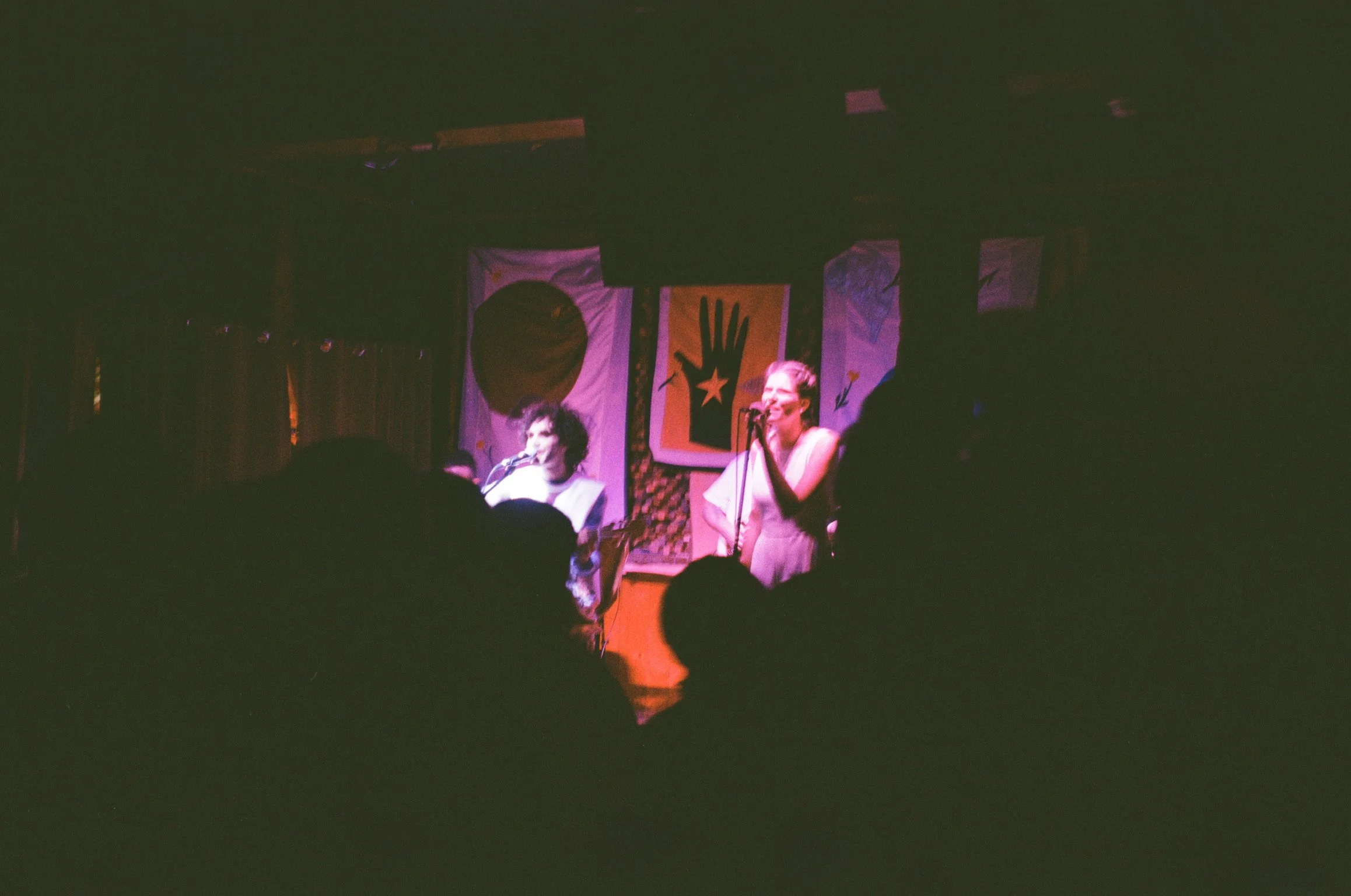 Two women perform on a dimly lit stage with colorful banners and artwork in the background, audience members' silhouettes are visible in the foreground.