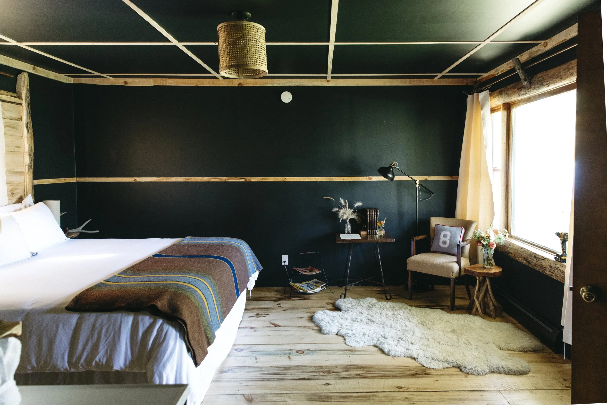 A cozy bedroom with a black accent wall, a bed with white linens and a multicolored striped blanket, wooden floors, a small side table with flowers, a chair with a cushion, an armchair, and a large window with curtains, natural light filling the room