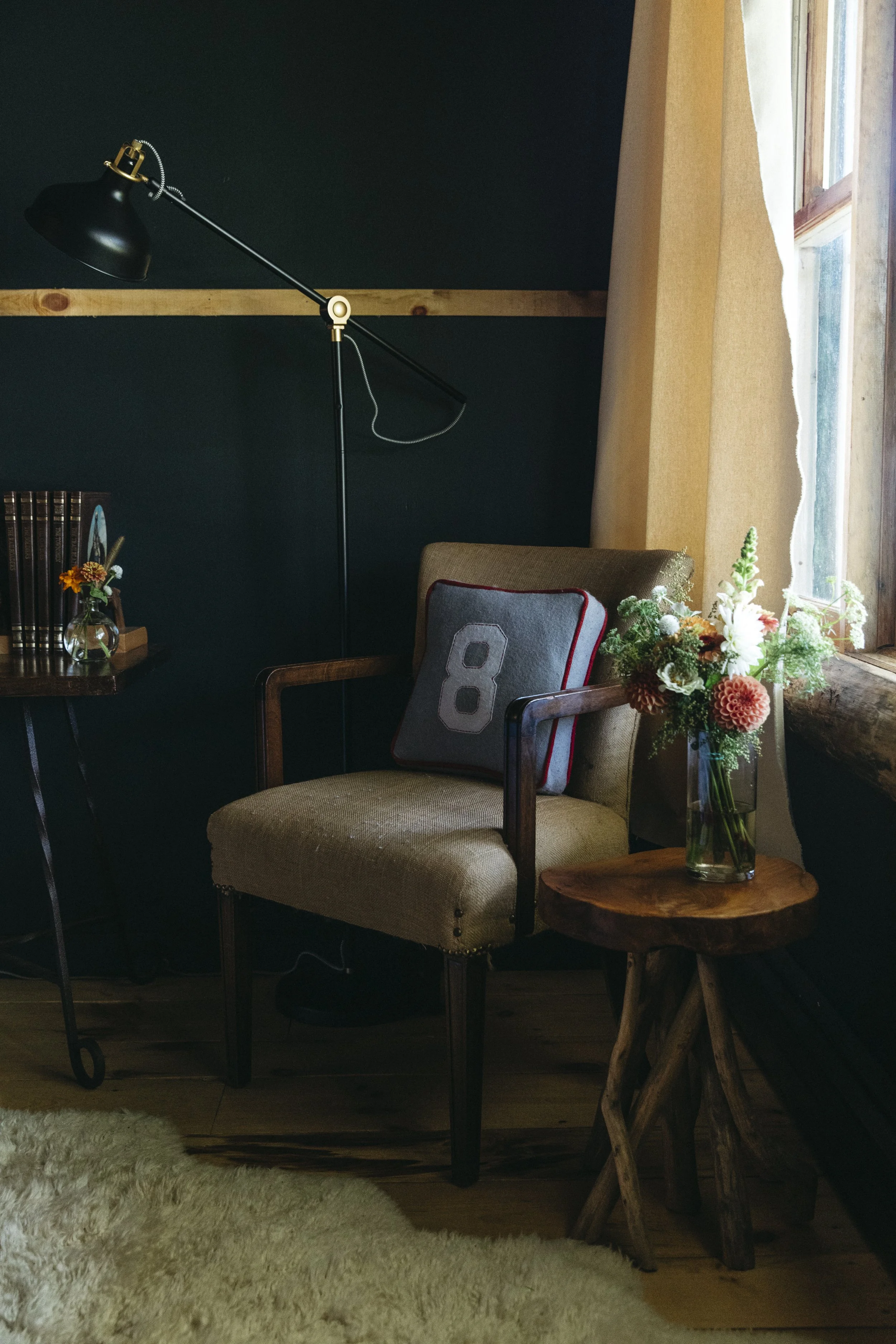 A cozy corner with a vintage beige armchair, a small round wooden side table with a glass vase of colorful flowers, a black wall-mounted adjustable lamp, a bookshelf with books and a small flower arrangement, and a window with curtains letting in nat