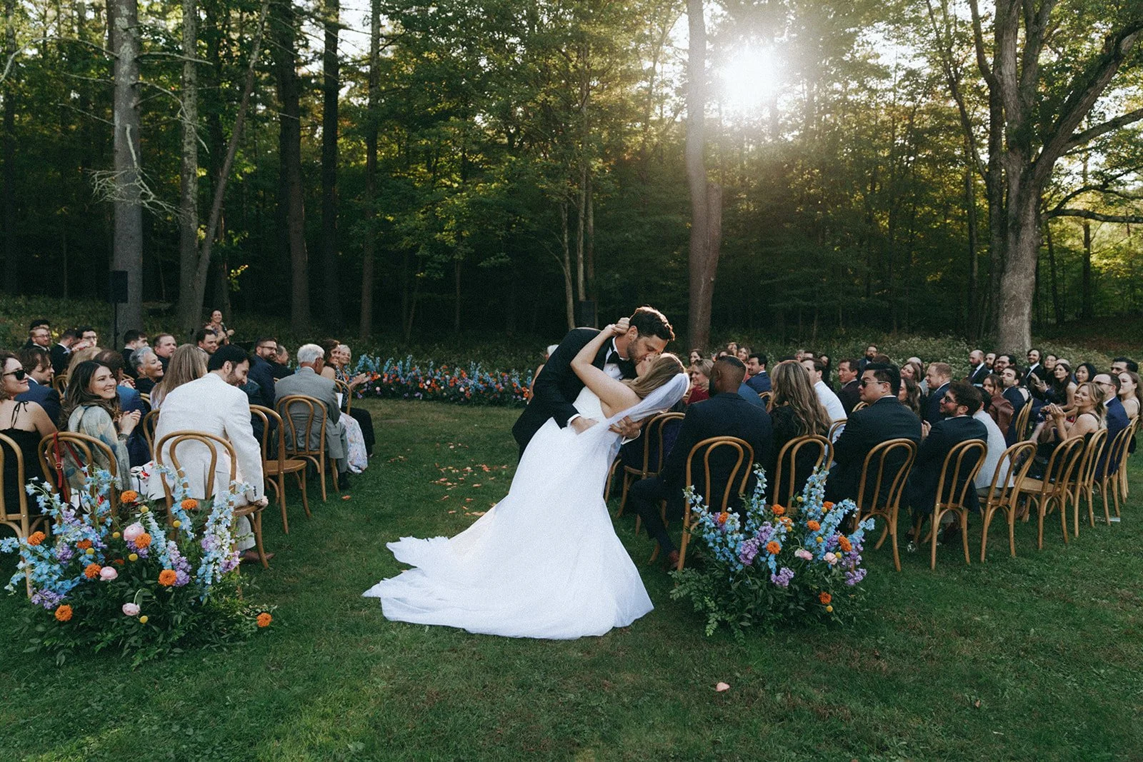 A bride and groom share a kiss during their outdoor wedding ceremony surrounded by seated guests and colorful floral arrangements, with a forest backdrop and sunlight filtering through trees.