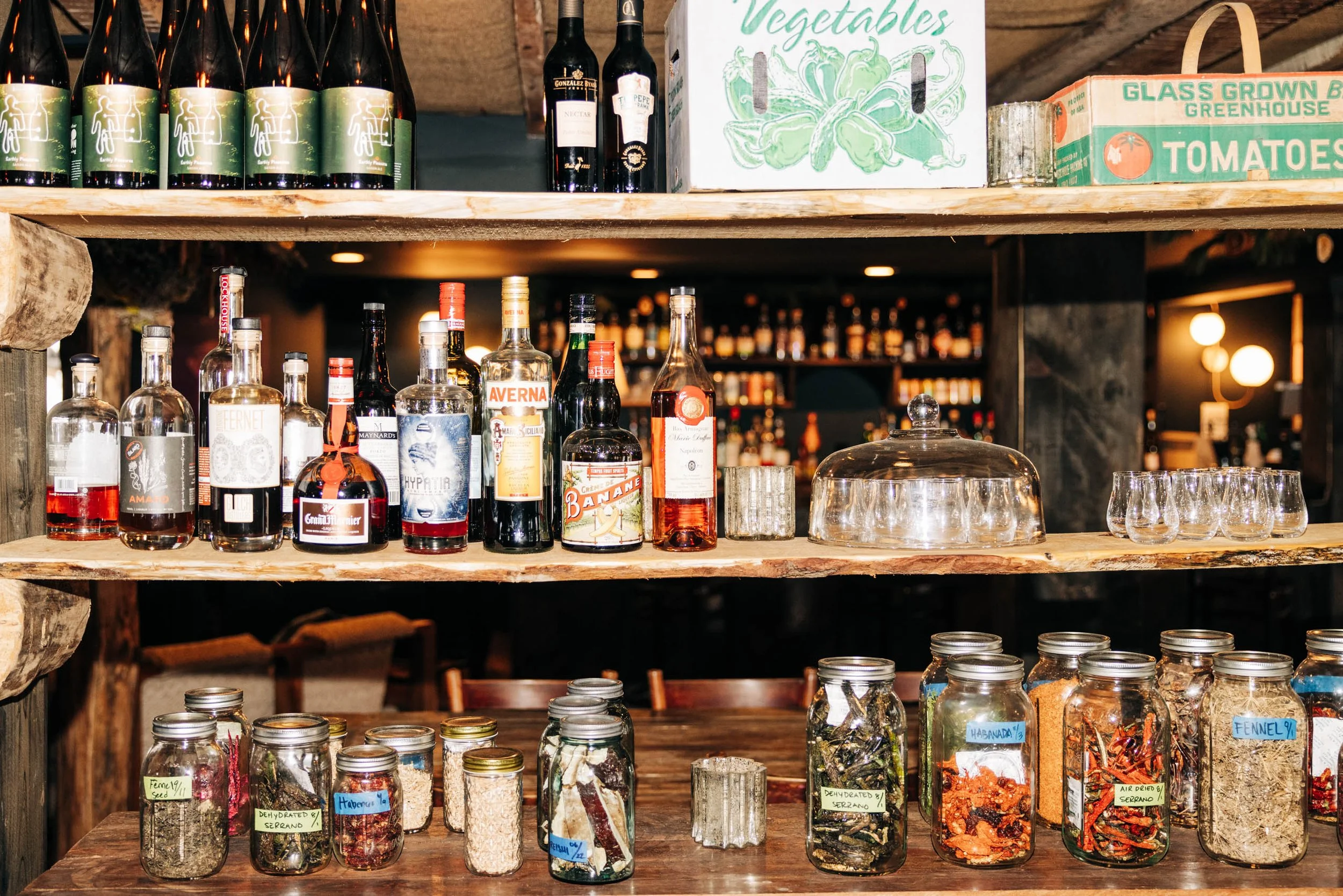 Shelves with various bottles of alcohol, jars of dried herbs and spices, and a sign labeled 'Vegetables' in the background.