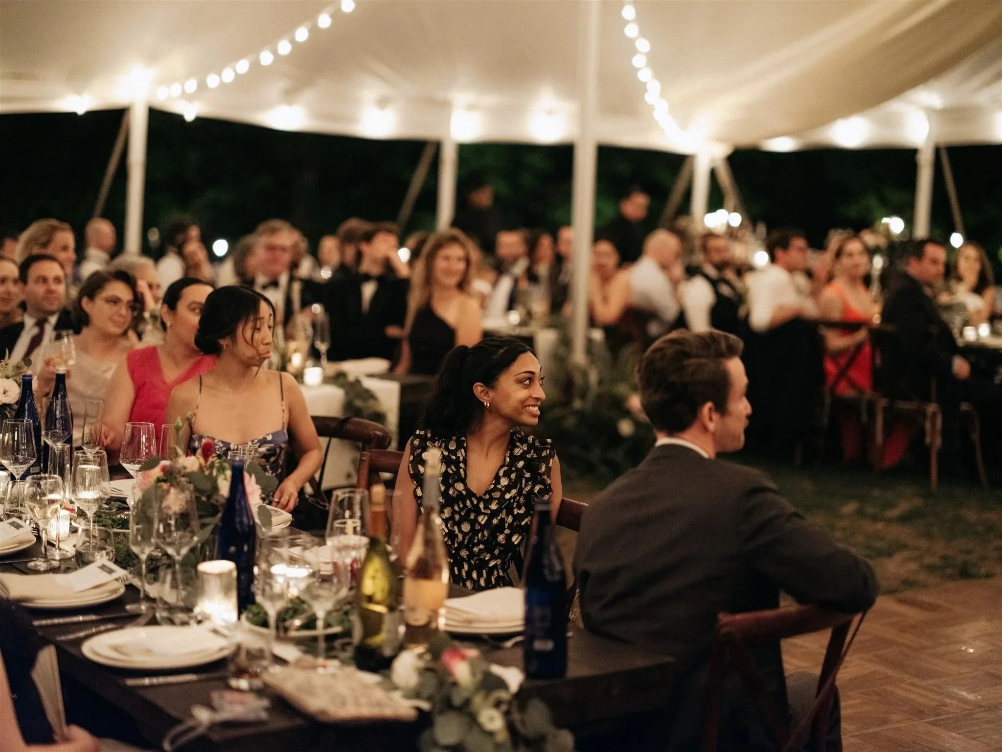 Guests seated at tables under a decorated tent at an evening event, with string lights overhead,  some smiling and enjoying the occasion.
