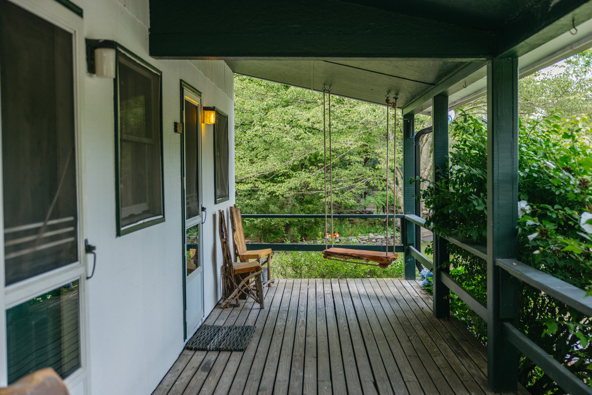 A porch with wooden flooring, green railings, two wooden chairs, a swing hanging from the ceiling, and outdoor greenery in the background.