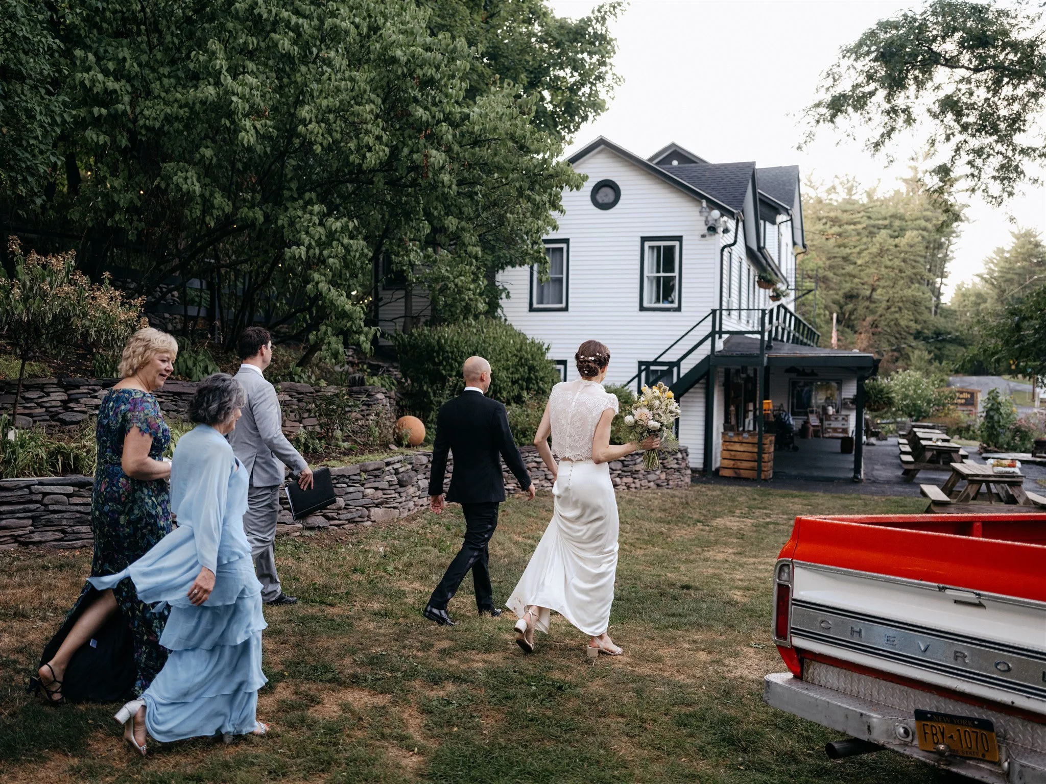 A bride in a white dress holding a bouquet walks towards a house with four guests following her, in a garden setting with trees and outdoor furniture.