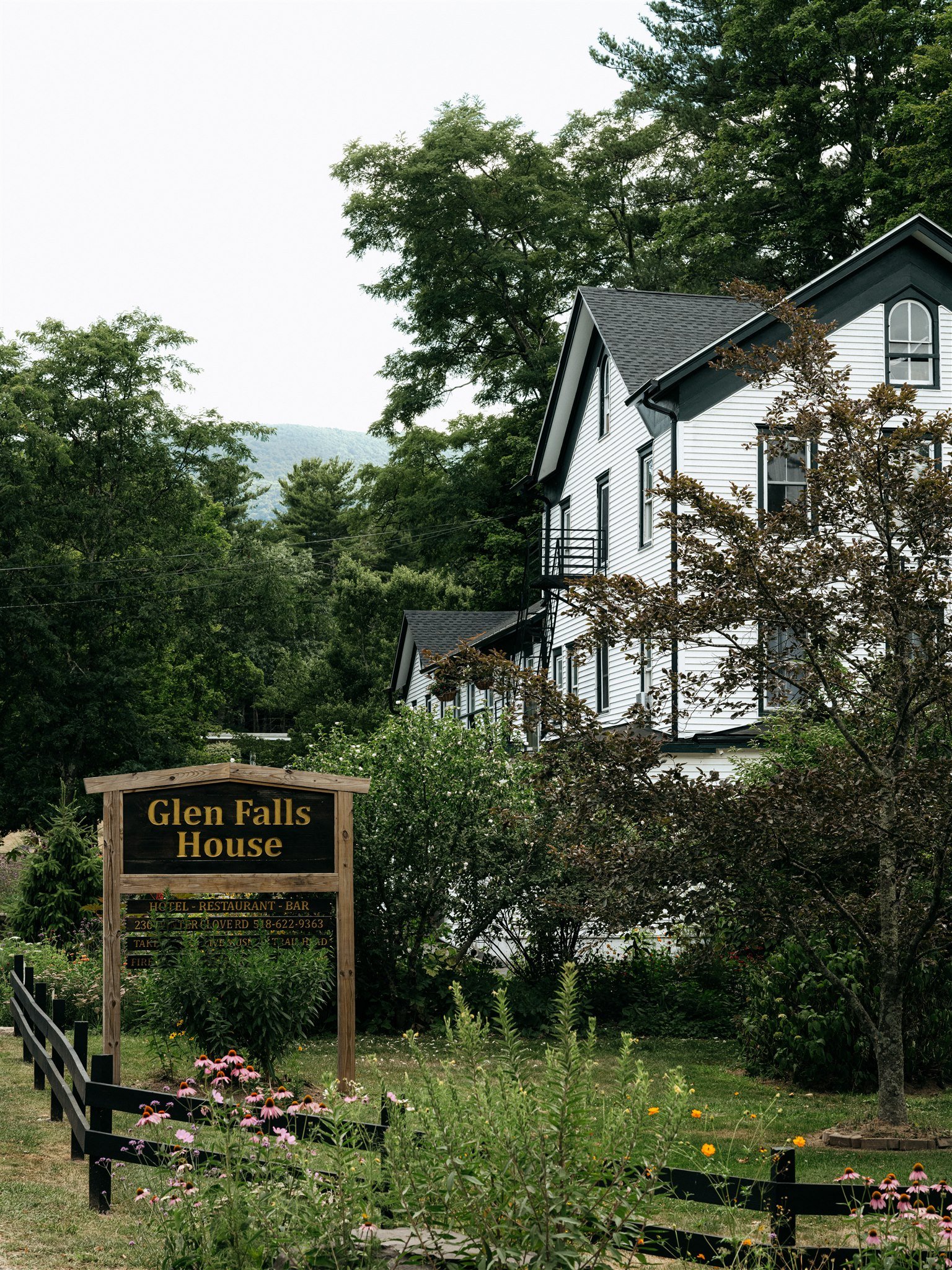 A large white house surrounded by trees and flowering plants with a wooden sign that reads 'Glen Falls House' in front.
