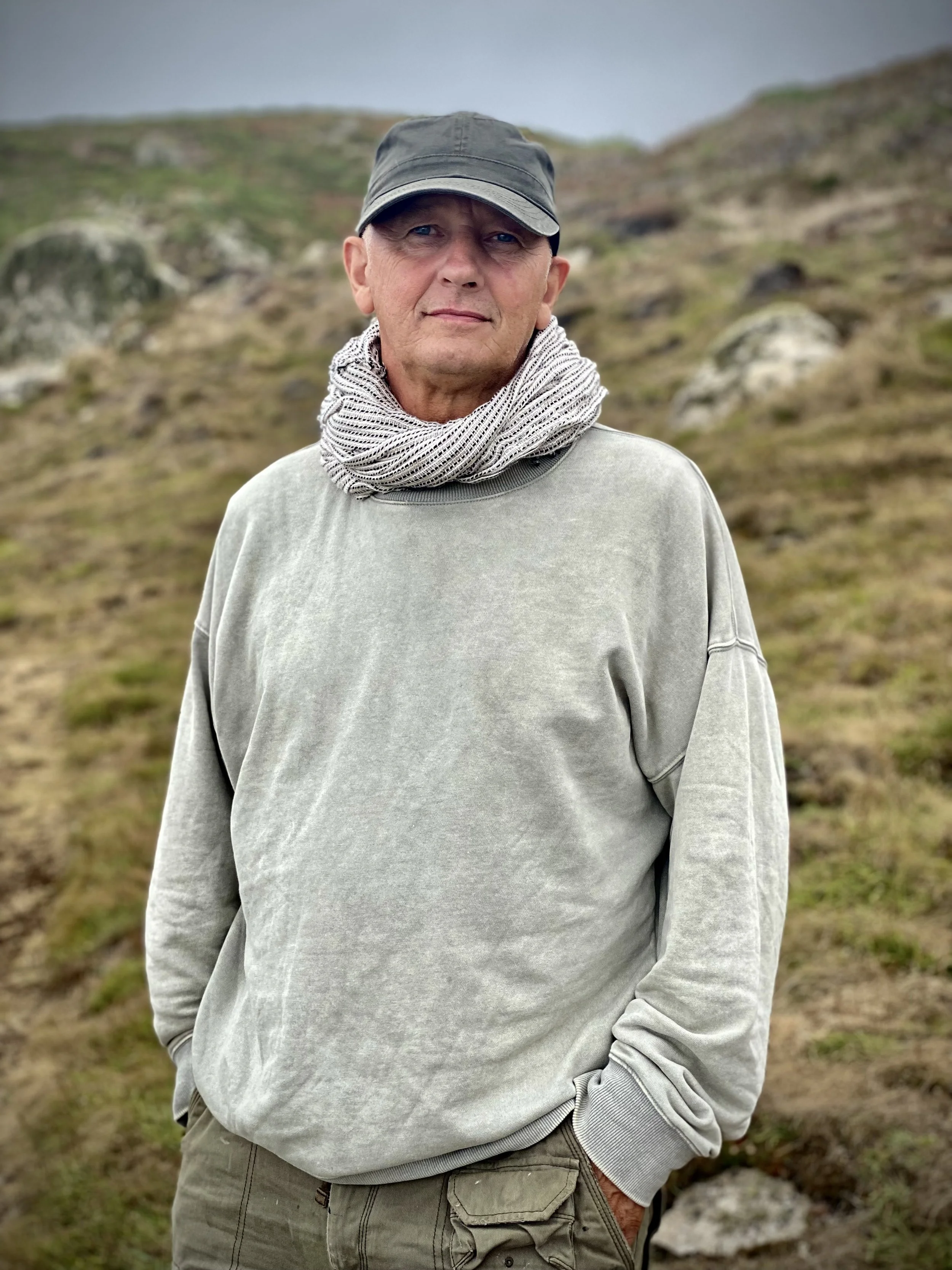 Lewes artist John Worth standing outdoors in a grassy, rocky landscape wearing a gray sweatshirt, beige pants, a gray hat, and a patterned scarf.