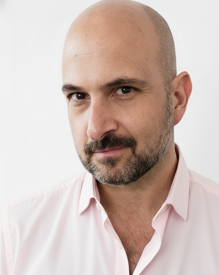 Headshot of a bald man with a beard wearing a light pink button-up shirt with a serious expression.