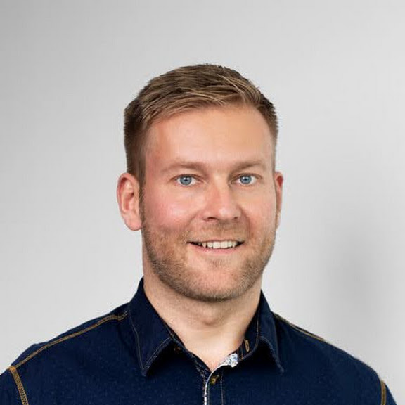 A smiling man with short blond hair and blue eyes wearing a dark blue shirt against a gray background.