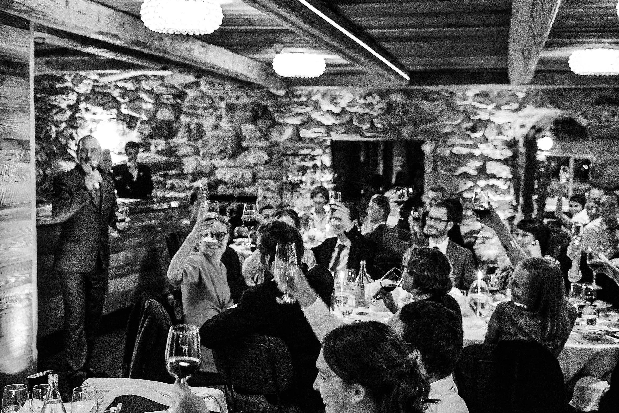 Groupe de personnes à un dîner, levant leurs verres de vin pour porter un toast dans une salle avec murs en pierre et plafond en bois.