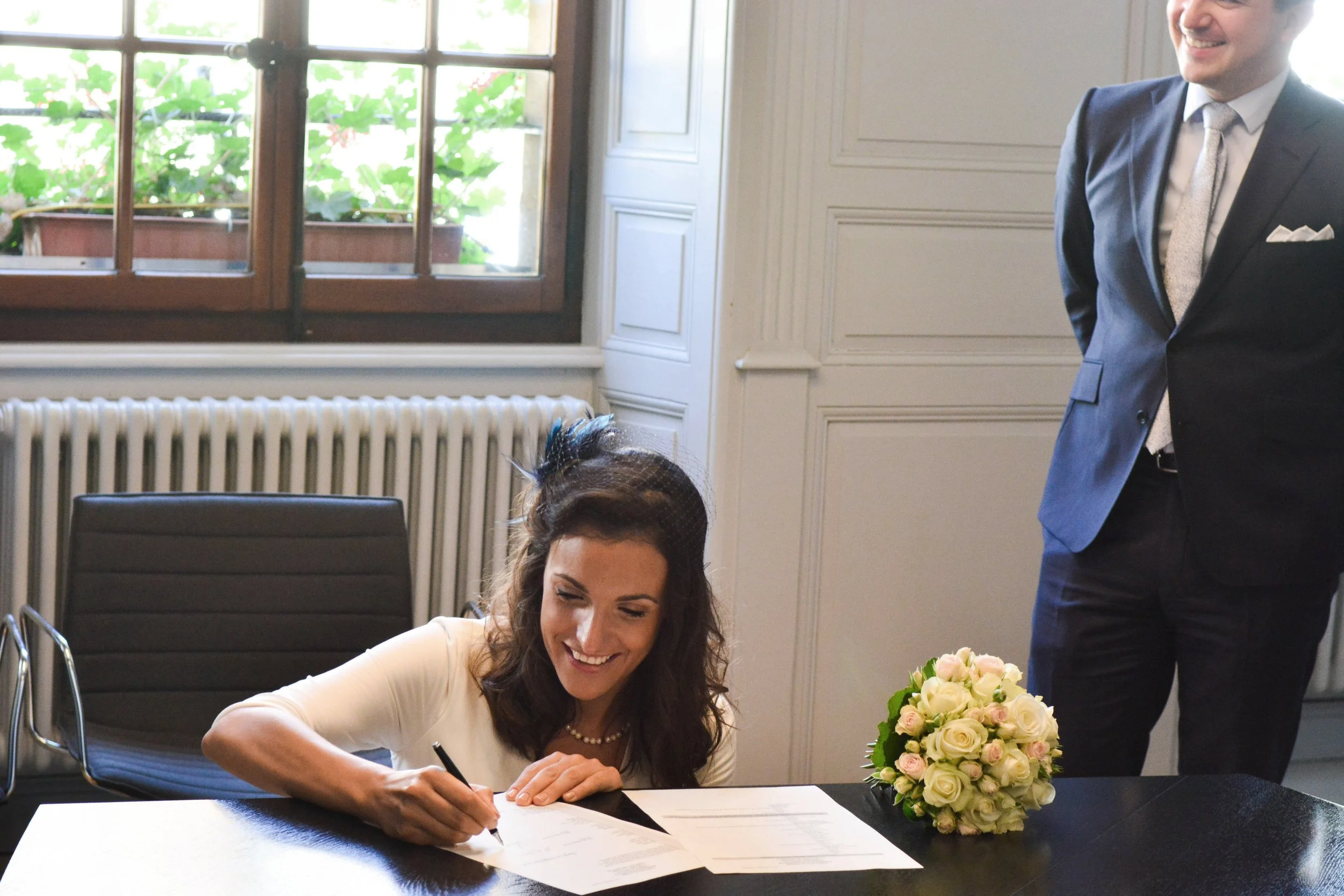 Une femme souriante signe un document sur une table noire avec un bouquet de fleurs blanches à côté, un homme en costume se tient debout derrière elle dans une pièce bien éclairée par une fenêtre.