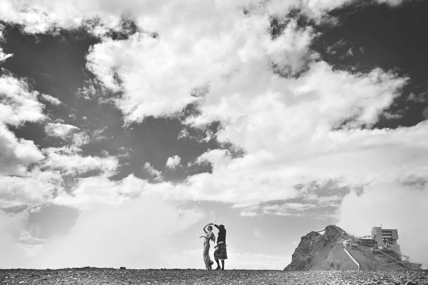 Deux personnes dansent sur un pierrier avec une structure rocheuse et un bâtiment en arrière-plan, sous un ciel nuageux.