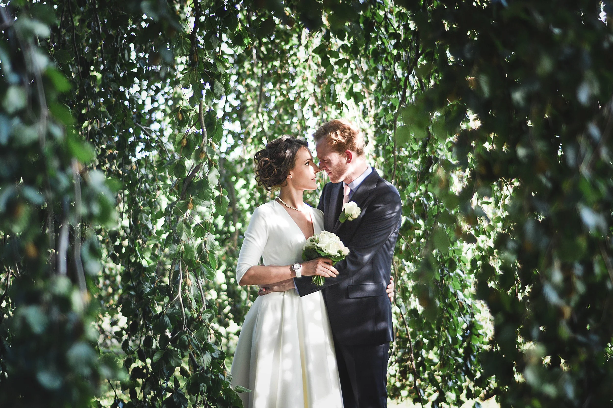 Un couple de mariés, un homme en costume noir et une femme en robe blanche, se tient dans un environnement naturel entouré de feuillage vert, se regardant tendrement, la femme tient un bouquet de fleurs blanches.