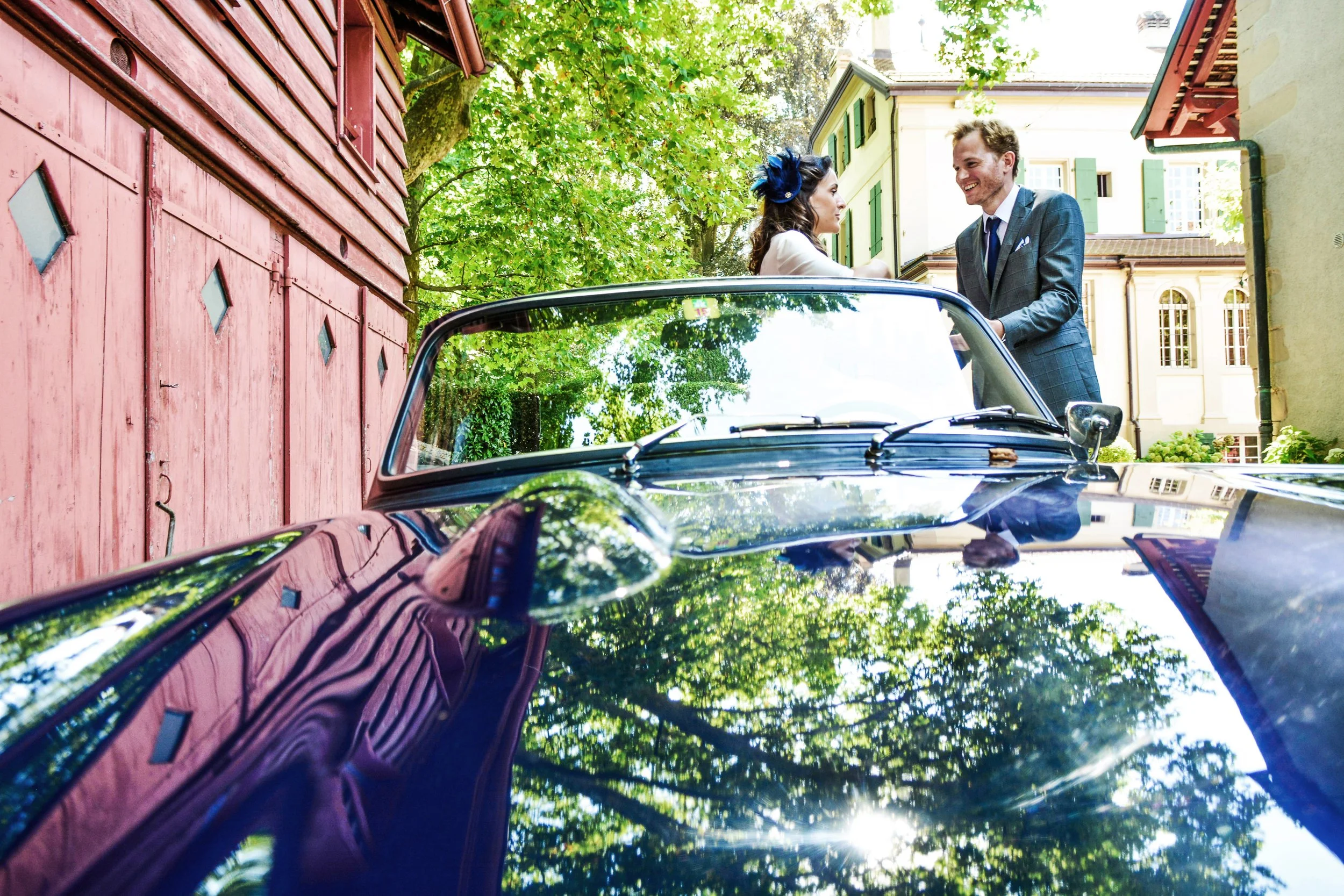 Un couple chic discutant à côté d'une voiture de collection noire dans un jardin verdoyant avec des bâtiments anciens en arrière-plan.