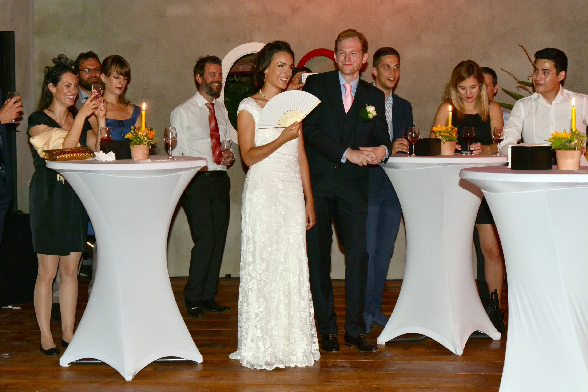 Un groupe de personnes lors d'une cérémonie de mariage, avec la mariée en robe blanche tenant un éventail blanc et le marié en costume noir avec une rose, entourés d'amis et de famille qui prennent des photos et regardent la scène.