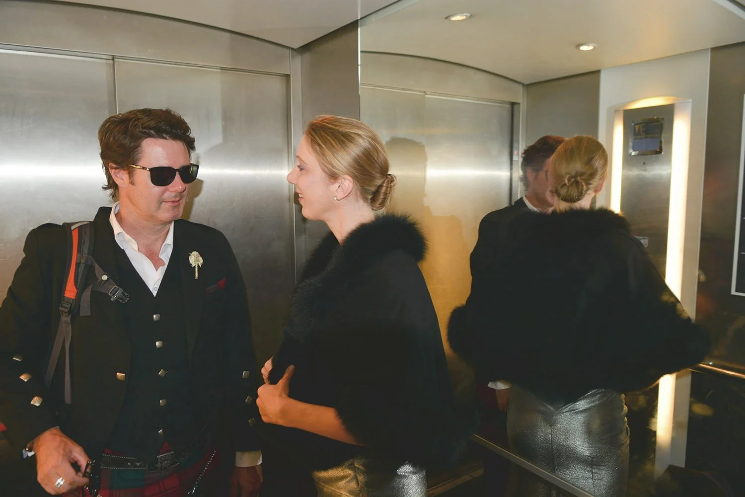 Homme avec lunettes de soleil et femme parlant dans un ascenseur, portant des manteaux noirs et des vêtements métalliques, arrière-plan en acier.