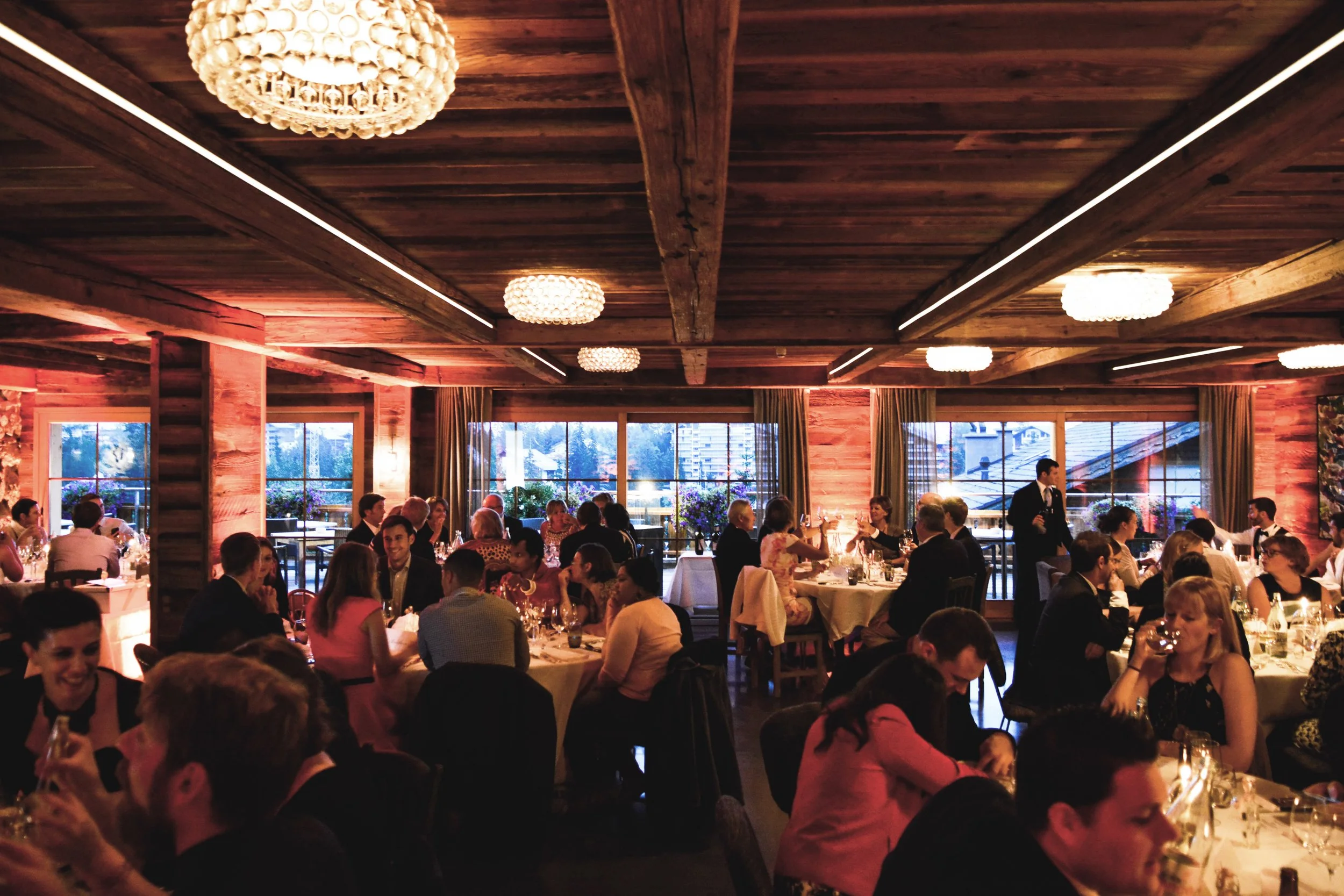 Une salle de banquet en bois avec plusieurs tables occupées par des personnes en train de manger et de parler, éclairée doucement par des chandeliers