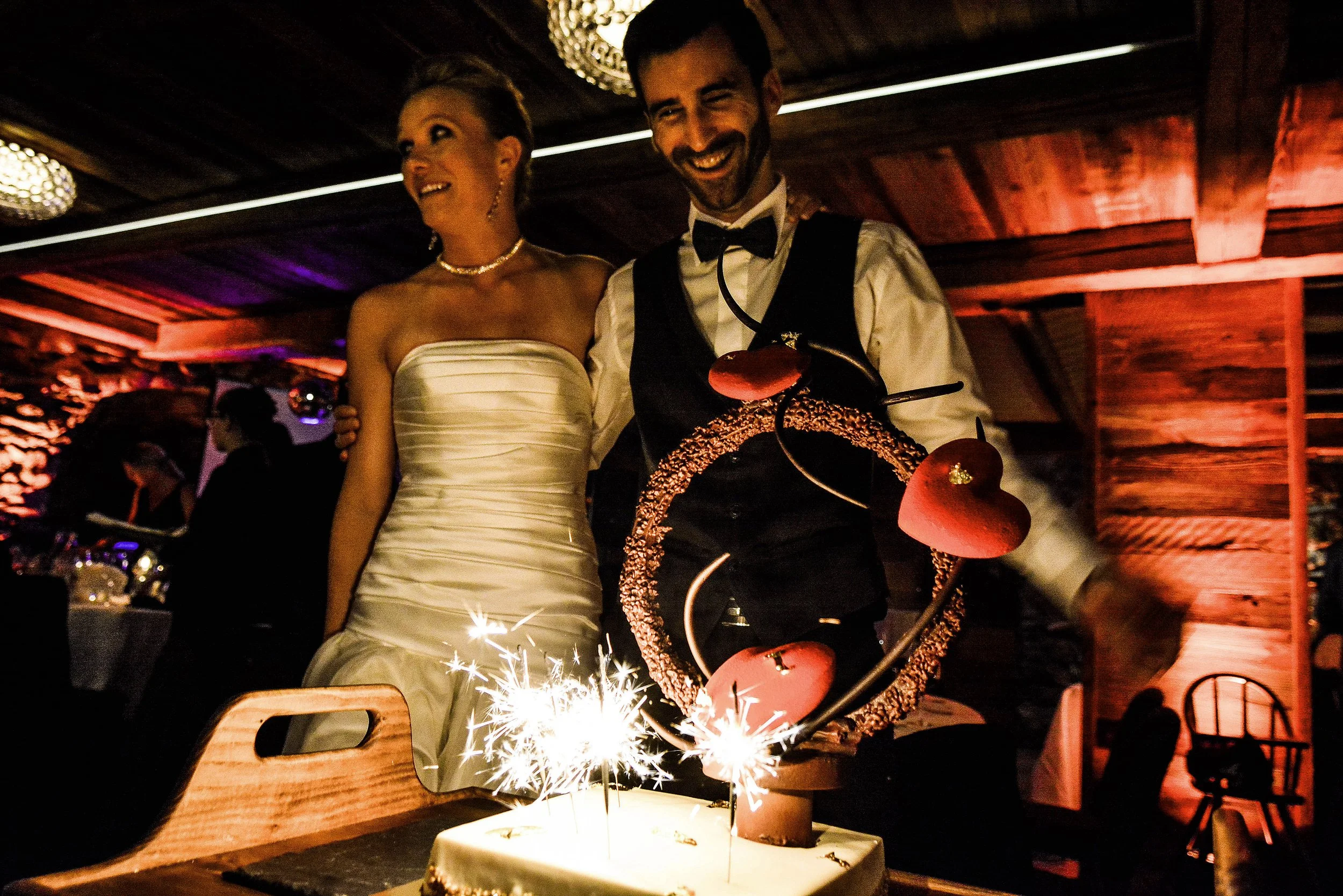 Un couple en tenue de mariage célèbre un anniversaire avec un gâteau décoré de magnolias, des bougies et des feux d'artifice dans un cadre chaleureux en bois.