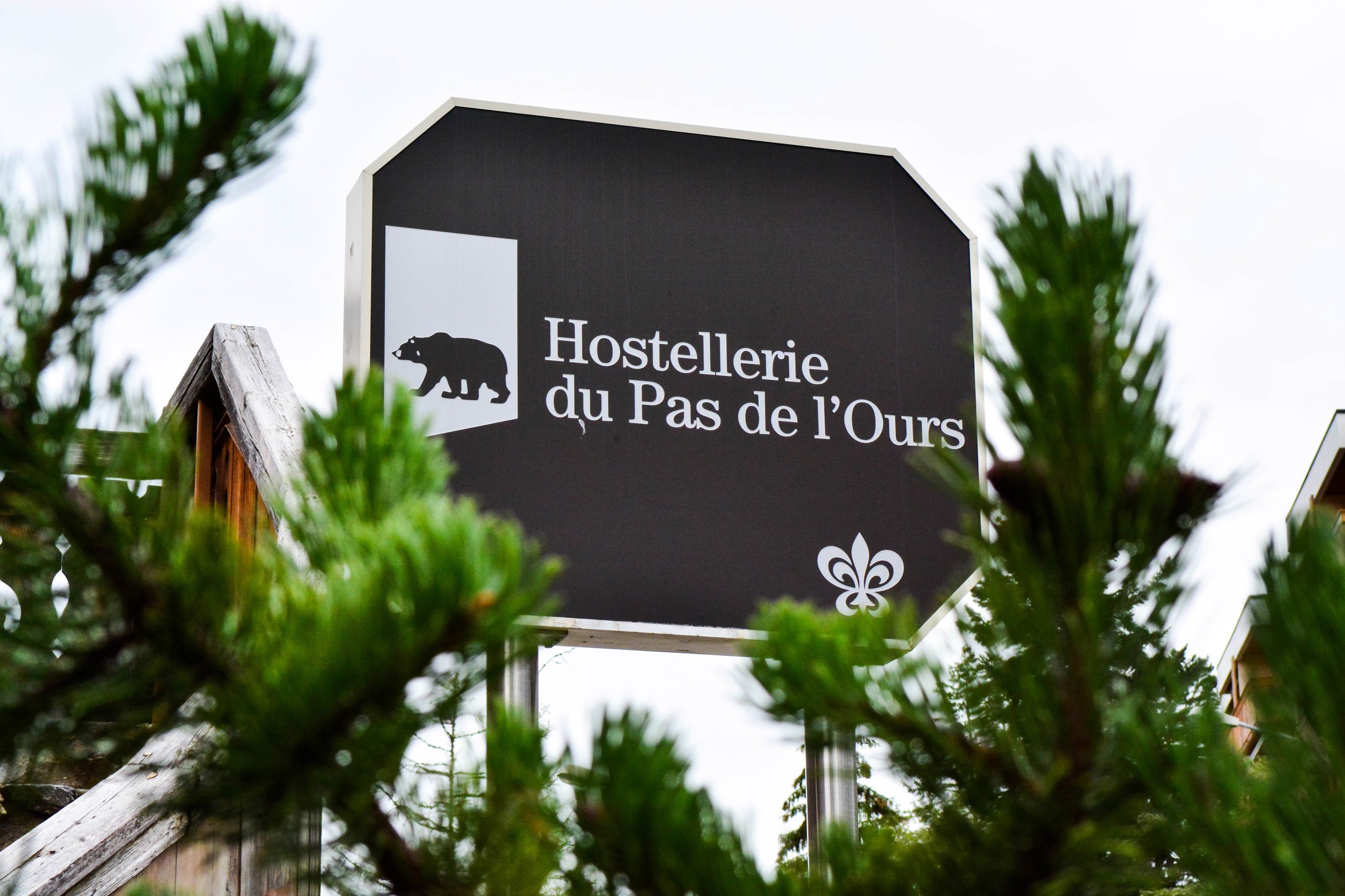 Un panneau noir indique « Hostellerie du Pas de l'Ours » avec une icône de ours noir et une fleur de lys. Le panneau est partiellement entouré par des branches d'un arbre vert.